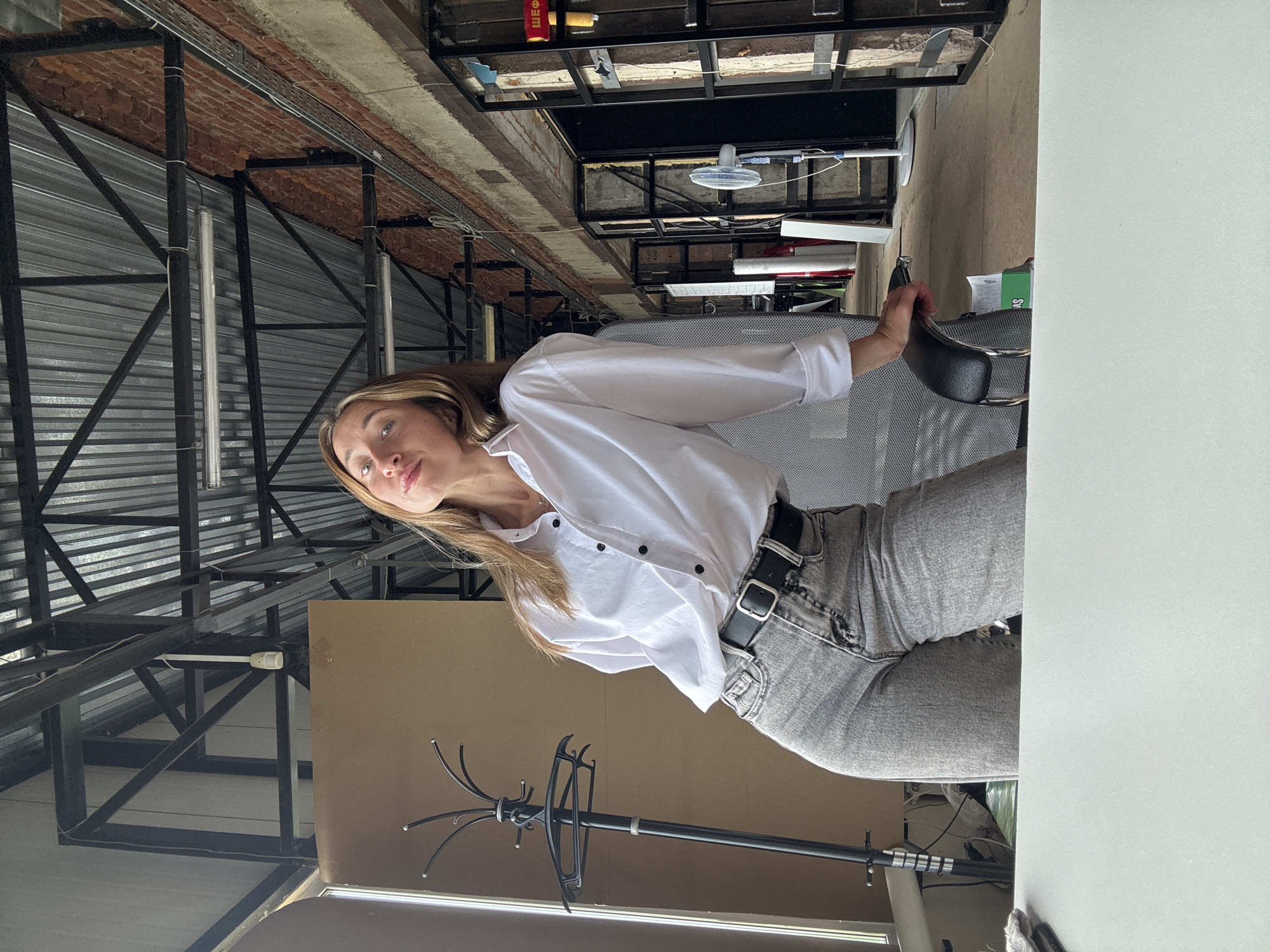 The image shows a young woman posing in an industrial-style open-plan space, possibly a workshop or studio, located in Chaikovsky, Russia. The woman is wearing a white button-down shirt and gray jeans, with a black belt. She is leaning on a black office chair, with her right arm resting on its armrest. Her left hand is behind her back. The background features exposed brick walls, metal beams supporting a corrugated metal roof, and several doorways leading to other areas. A coat rack is visible to the left of the woman, and a white desk is in the foreground. A white standing fan and a red hammer are visible further in the background. The lighting suggests it is daytime.