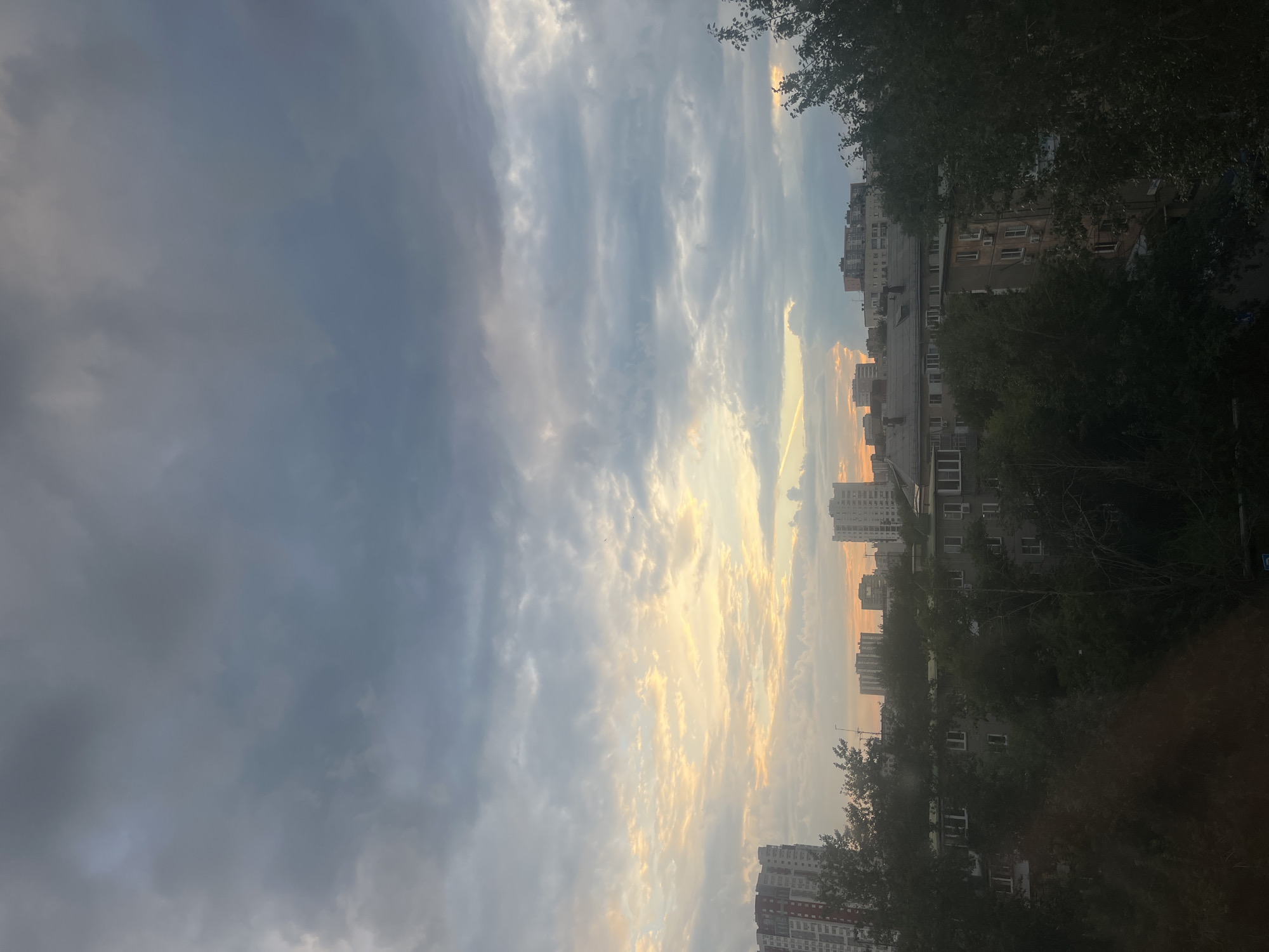 The image captures a dusky sky over a residential area. The sky is a dramatic canvas of clouds, with streaks of golden light from a setting sun piercing through the greyer, more ominous clouds. The contrast between the warm sunlight and the cool tones of the clouds suggests a transition in weather or time of day.
Below the sky, buildings are silhouetted, interspersed with dark, leafy trees. These appear to be apartment blocks, typical of urban or suburban landscapes. Their architecture suggests a location in Russia, consistent with the provided context of Chelyabinsk. The trees, with their dense foliage, partially obscure the buildings, adding depth and texture to the scene.
There is no discernible activity or event taking place; the image seems to be a still capture of the urban environment during twilight. The overall mood is one of quiet observation, with the dramatic sky providing a focal point. The time of day is clearly evening, as indicated by the quality of the light. No text is visible in the image.