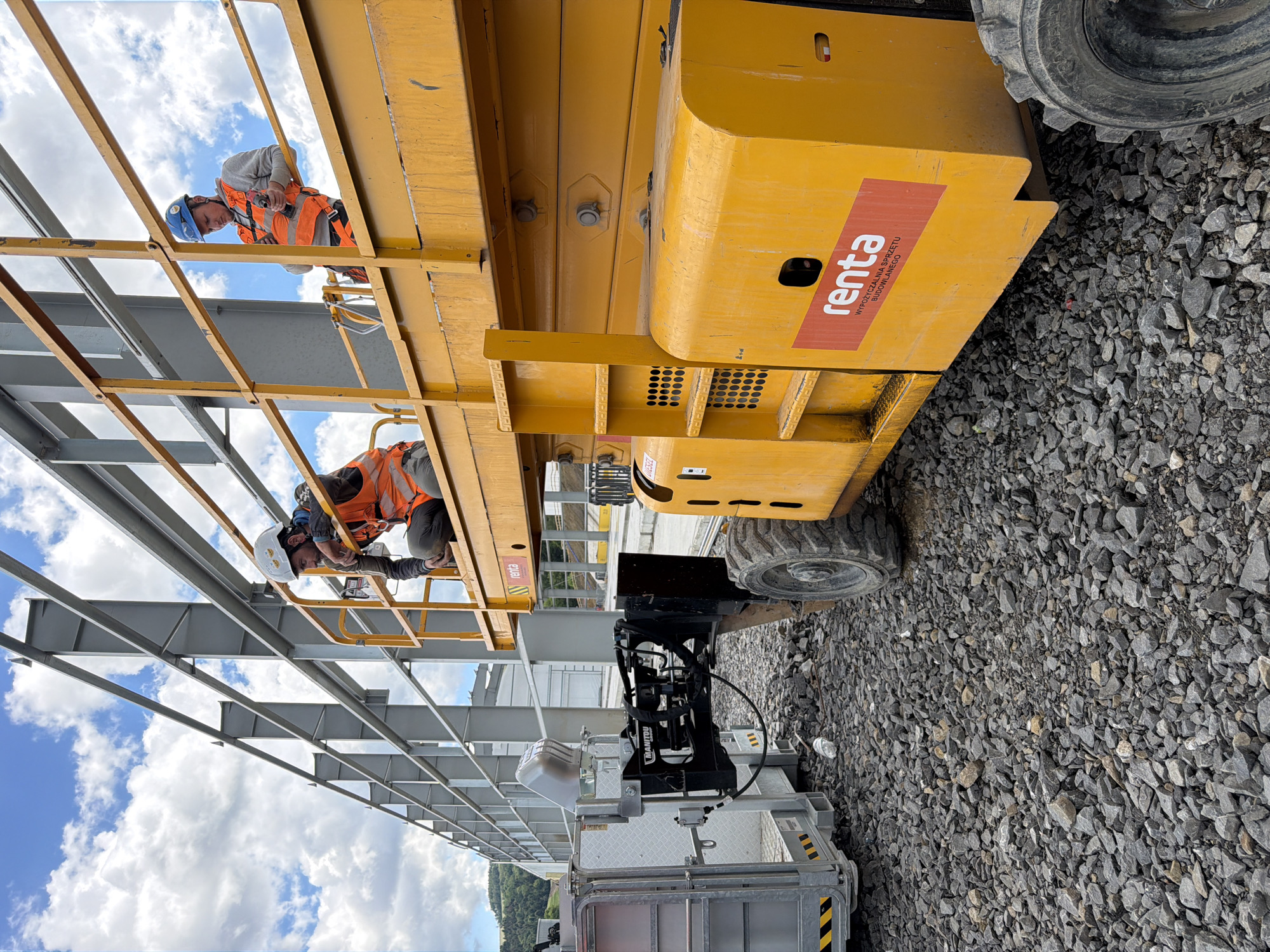 Two construction workers are on a yellow scissor lift, with one worker kneeling and looking down, while the other stands and operates a power tool. The scissor lift is positioned on a surface covered in gravel, adjacent to a large metal construction structure. In the background, there are hills and a cloudy sky. The logo "renta" is visible on the side of the scissor lift, indicating it is a rental company. The scene is set in Janowice, Poland.