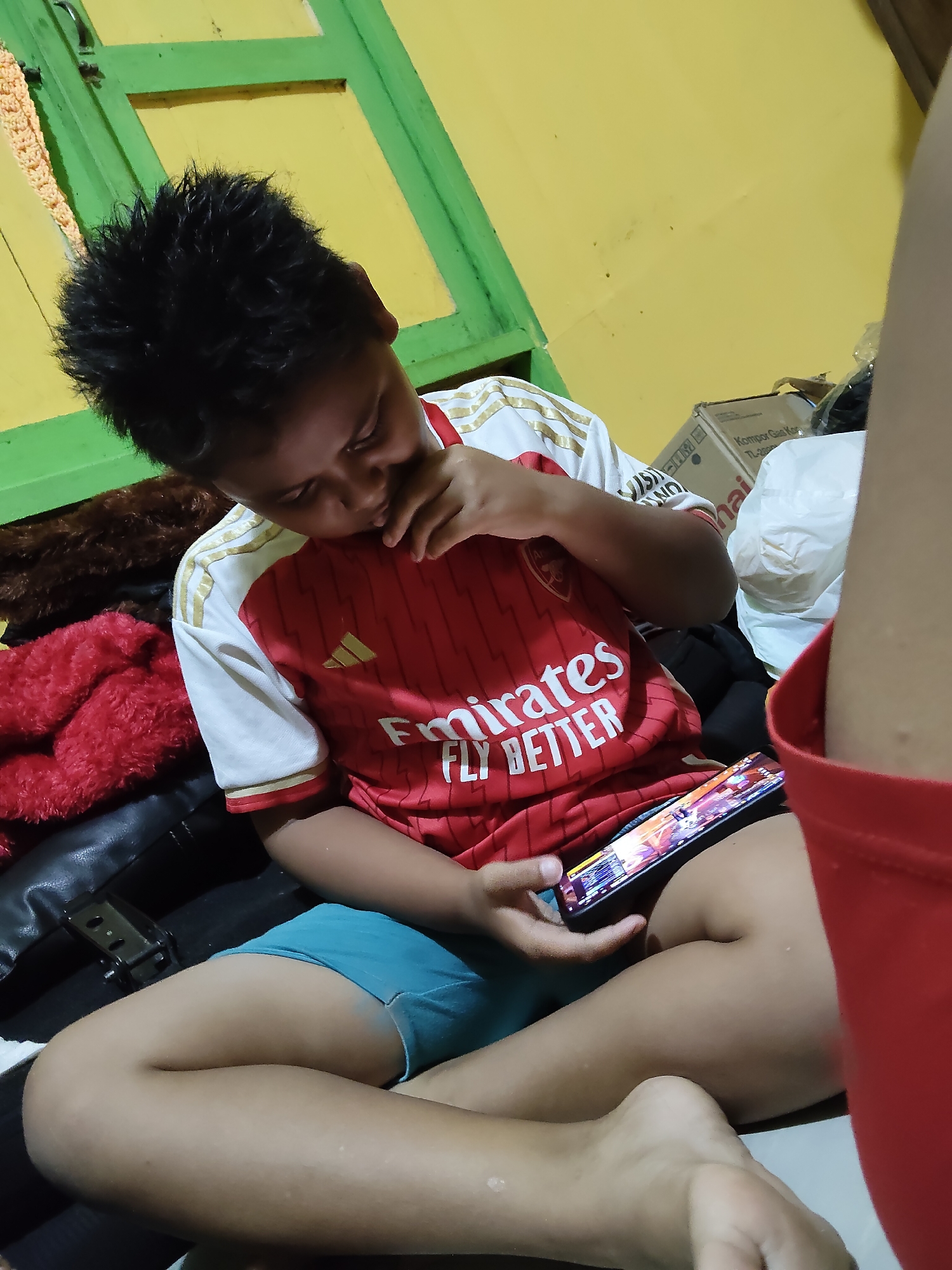 A young boy, likely of Indonesian descent and aged between 8-12, is seated on a dark-colored surface, possibly a bed or sofa, in what appears to be a residential setting in Tanah Grogot, Indonesia. He is wearing a red and white short-sleeved jersey with gold trim, which has the "Emirates Fly Better" slogan printed on the front. The boy is holding a smartphone in his right hand, the screen of which displays a game interface. His left hand is near his face, with his fingers touching his mouth, suggesting he might be contemplating or engrossed in the game.
The environment is dimly lit, with a yellow wall visible in the background, painted with green trim around a window or door frame. Several objects are scattered around the scene, including a brown furry blanket, a red fluffy blanket, and a white plastic bag. A cardboard box with text, possibly a product label, is also present in the background. The boy is wearing light blue shorts. The overall impression is one of a casual, domestic scene where the boy is occupied with his mobile device.