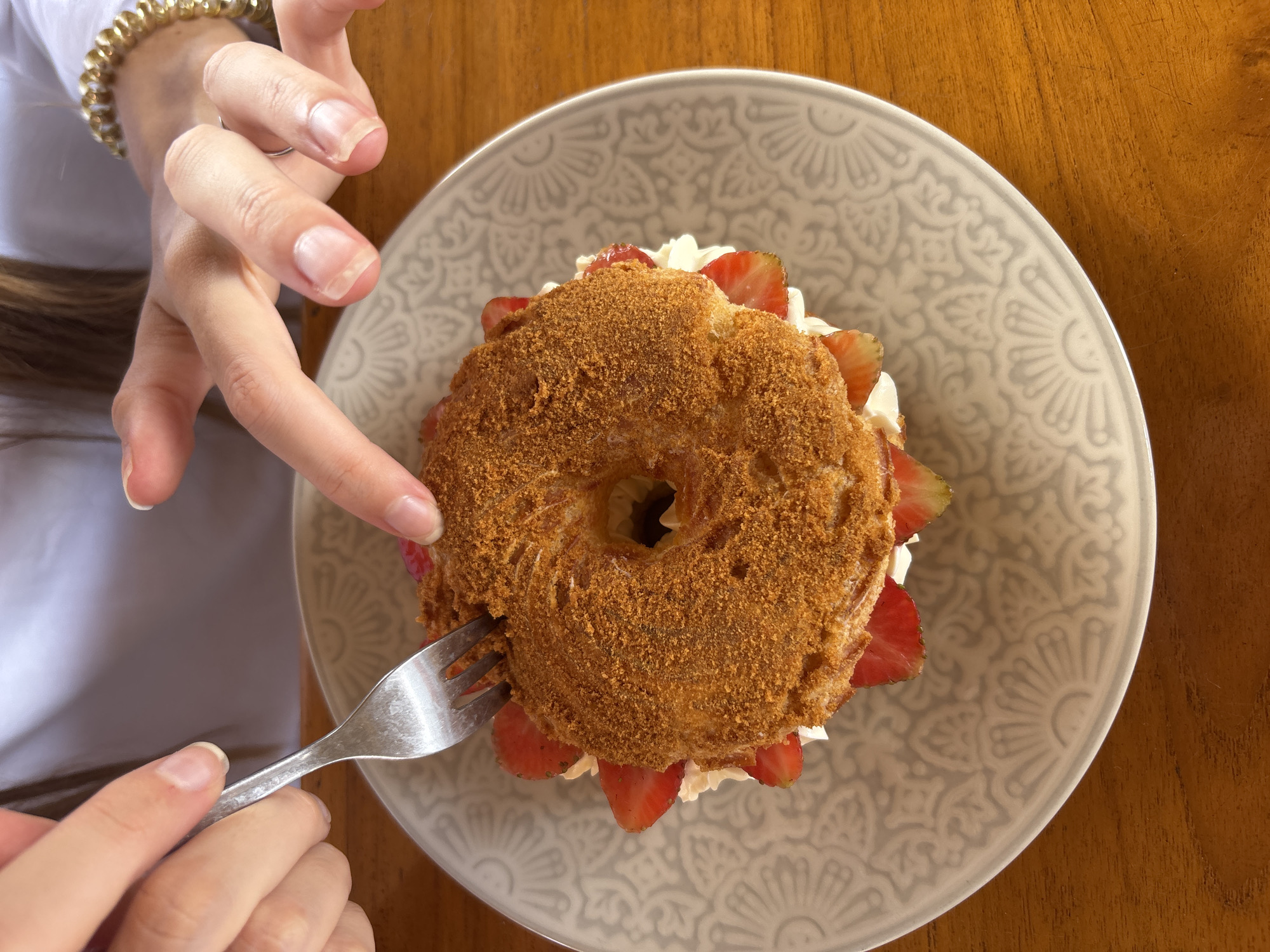 A close-up, overhead shot shows a person's hands about to eat a large, ring-shaped pastry filled with cream and strawberries. The pastry is covered in a brown, crumbly topping. The pastry sits on a decorative grey plate, which is placed on a wooden table. The person is using a silver fork to cut into the pastry. The person's left hand is holding the fork, while their right index finger is pointing at the pastry. The background is blurred, showing a white shirt and a golden bracelet on the person's wrist. The scene is likely a cafe or restaurant in Kuta Selatan, Indonesia, given the lighting and the wooden table common in such establishments.