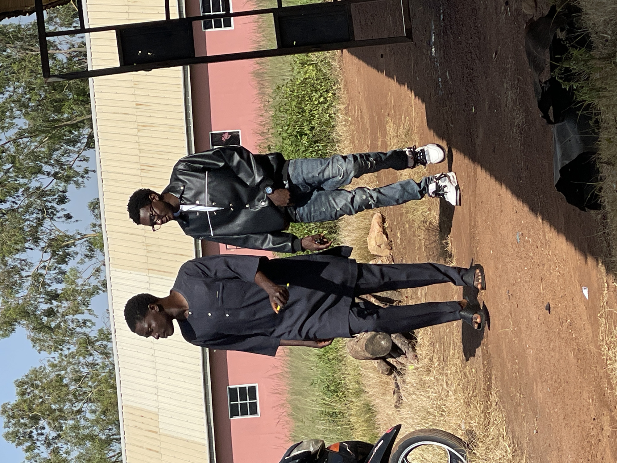 Two young men walk side-by-side on a dirt path in Durumi, Nigeria. The man on the left wears a dark blue traditional tunic and trousers, with black sandals. He holds a yellow object in his right hand and is looking to his left. The man on the right wears a black leather jacket over a white t-shirt, distressed denim jeans, and white sneakers. He is looking forward and to his right. Behind them, a pink building with a corrugated metal roof stands, and trees with green foliage are visible against a clear blue sky. A motorcycle is partially visible on the left side of the image, and a metal gate structure is on the right. The ground is dry and dusty, with scattered dry grass and a few small rocks.