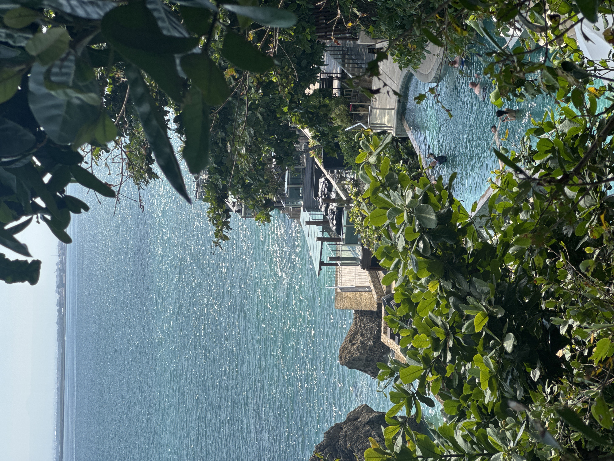The image shows a coastal scene in Kuta Selatan, Indonesia, viewed through foliage. The ocean is visible, sparkling in the sunlight. A cliff-side structure, possibly a hotel or resort, is present with wooden decks and terraces. A pool is seen with several people swimming in it.
