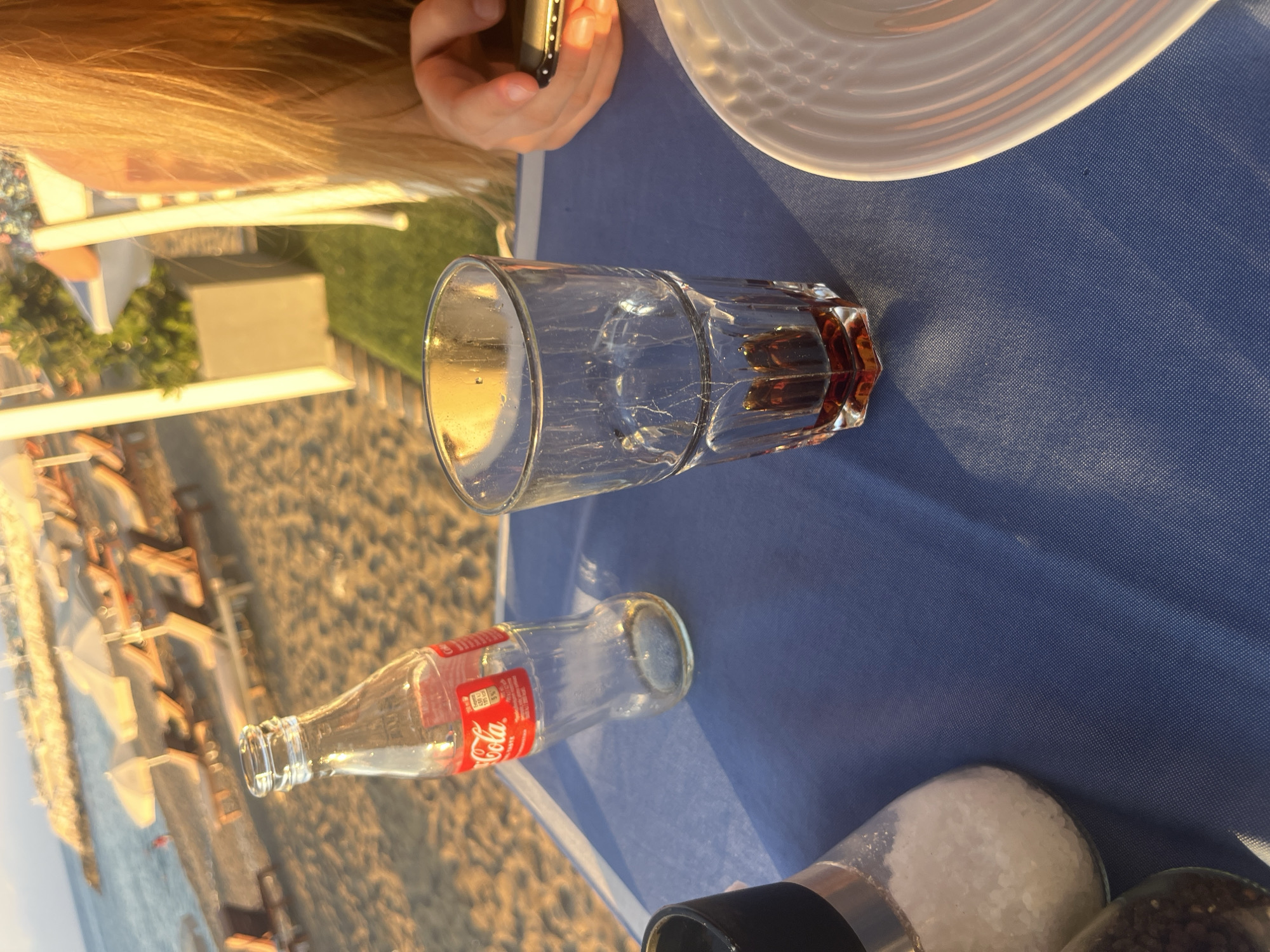 A close-up outdoor shot captures a table set on a beach in Polis Chrysochous, Cyprus. A blue tablecloth covers the table, with a salt shaker on the left and a partially filled glass of dark liquid and an empty glass Coca-Cola bottle to its right. In the background, lounge chairs and umbrellas dot the sandy beach, and the sea is visible under a warm, golden light, suggesting late afternoon. A person's hand, holding a mobile phone, is visible in the upper right quadrant of the image.