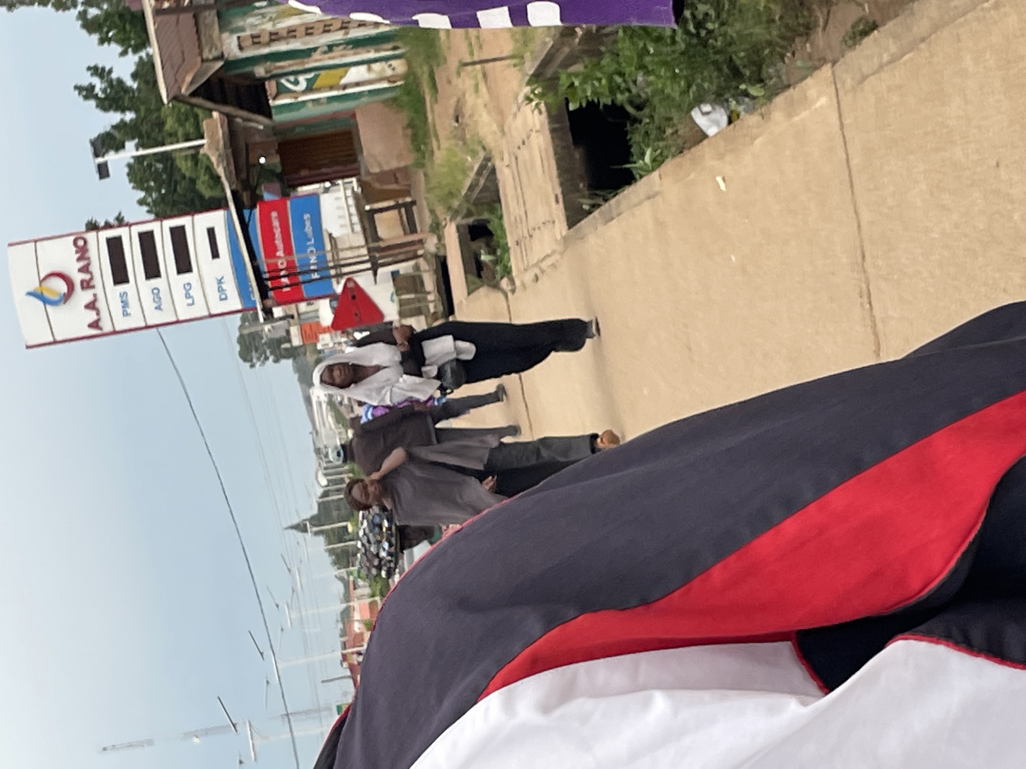 The image captures a street scene in Jos, Nigeria, likely on a cloudy day. In the foreground, a person's jacket, with red, white, and dark blue panels, is prominently visible. Further down the concrete sidewalk, two women are walking. The woman on the left wears a grey shawl over dark clothing and is speaking on her phone. The woman on the right is dressed in black pants and a white headscarf, carrying a black bag. Behind them, a man in a brown jacket walks away from the camera, and another person wearing a blue and purple garment is partially visible on the far right.
Dominating the background is a tall, white A.A. RANO gas station sign displaying fuel prices: PMS, AGO, LPG, and DPK. Adjacent to the sign is a small building with a corrugated iron roof and painted walls. A drainage channel with a concrete cover runs alongside the sidewalk, with overgrown vegetation and some litter visible within it. In the distance, a street with utility poles and what appears to be a collection of motorcycles can be seen. The overall environment suggests an urban setting with active pedestrian and vehicular traffic.