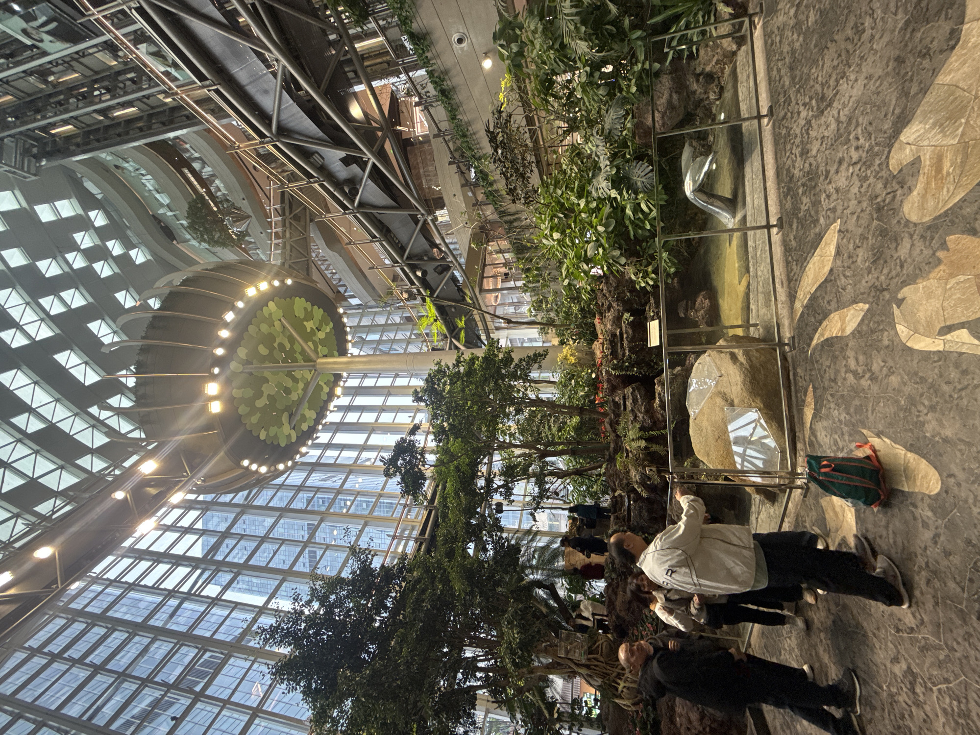 The image displays the interior of a multi-story atrium or lobby, identified as being in Liangjiang, China. The space is characterized by a towering glass ceiling with a geometric grid pattern and extensive glass curtain walls that reveal reflections of external high-rise buildings. A large, central indoor garden occupies the ground level, featuring mature trees, diverse green foliage, and exposed rock formations. Within this garden, a shallow water body contains a metallic, silver sculpture resembling a mermaid or fish tail partially submerged. A clear barrier with metal posts separates a viewing area from the water feature.
Suspended from the ceiling is a large, circular, illuminated canopy structure, abstractly resembling a stylized tree with green leaf-like elements. The architecture includes multiple curved balconies with glass railings at various levels, connected by visible escalators or walkways and supported by exposed steel trusses.
In the foreground, a group of approximately five adults stands on a patterned floor. One male, wearing a light-colored jacket, dark pants, and sneakers, leans forward, observing the water feature through the clear barrier. A green and orange backpack rests on the floor near his feet. To his left, another male in a dark jacket stands facing the garden. Other individuals are partially visible behind this group and scattered within the garden landscape and on upper levels of the atrium, appearing to observe the environment or transit through the space. The scene integrates natural elements with modern, large-scale architectural design.