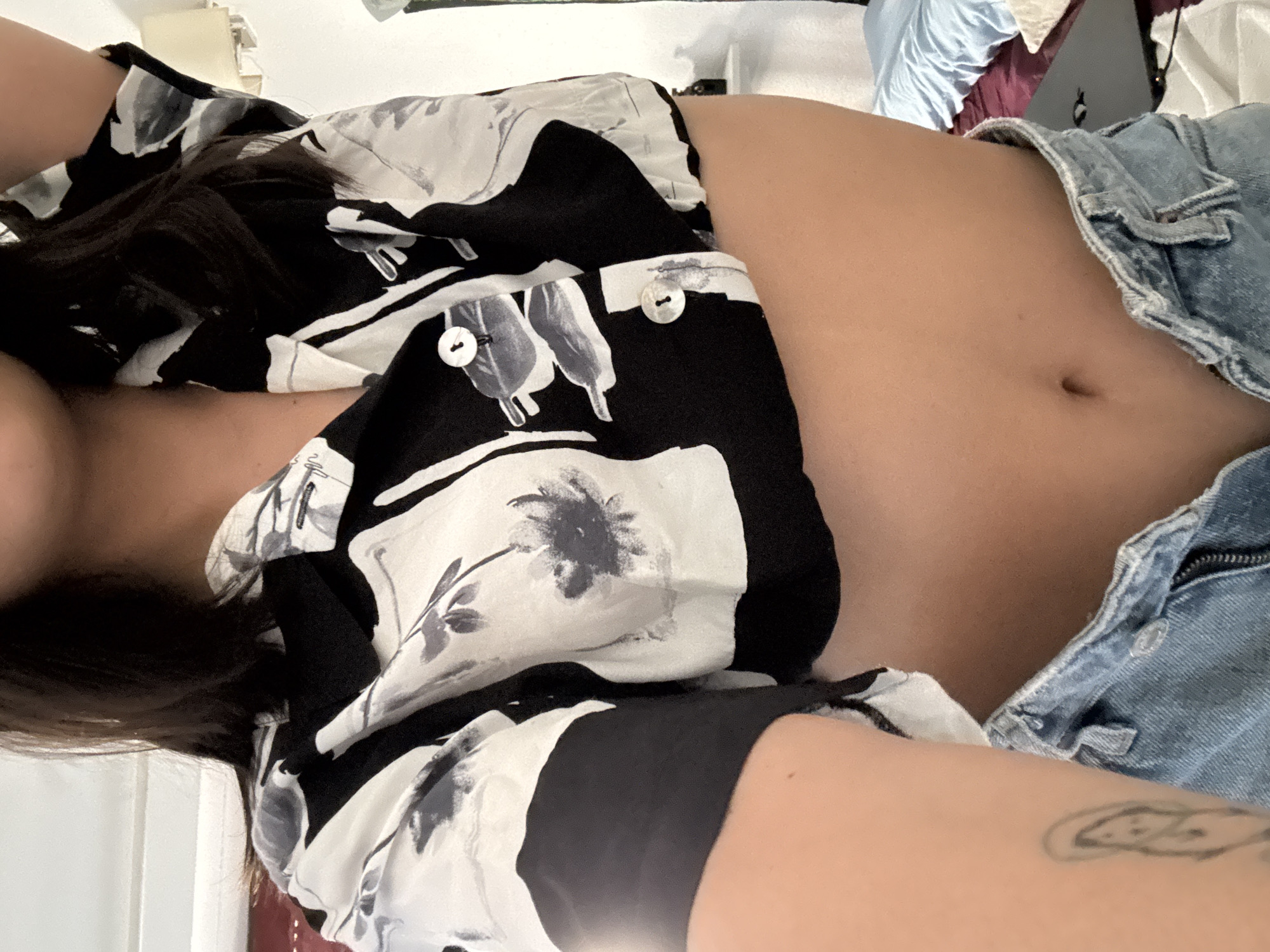 A person, whose face is not visible, is shown from the chest down, wearing a cropped black and white floral print shirt and light blue denim shorts. The shirt has short sleeves and a button-up front, with two visible white buttons. The denim shorts are unbuttoned and partially unzipped, revealing the person's midriff. A tattoo of a simple figure is visible on the person's left arm. The background appears to be an indoor setting, possibly a bedroom, with a bed, pillows, and a lamp visible. The lighting suggests it is daytime. There is no indication of the location being Las Rozas de Madrid, Spain.