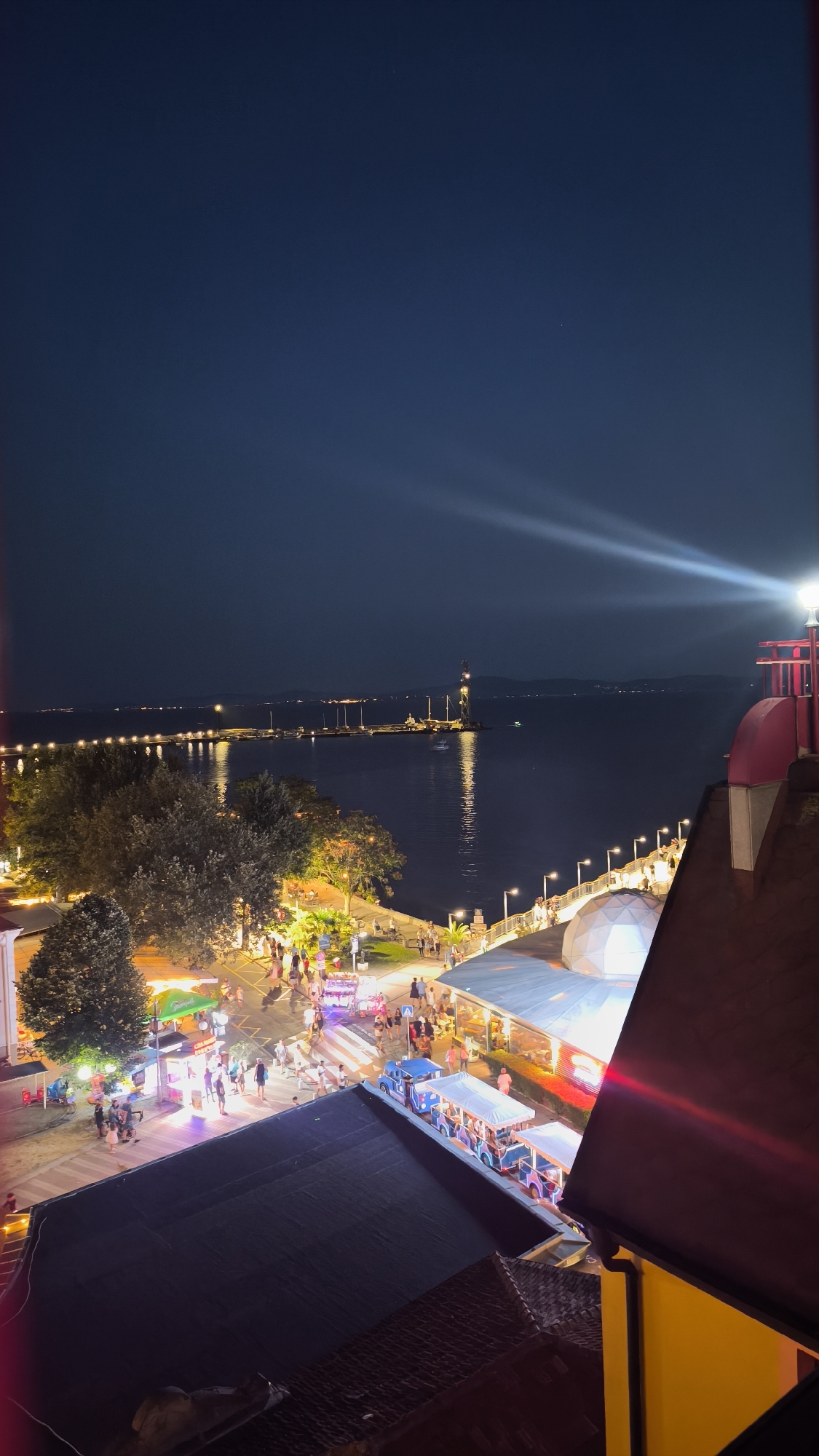 This image captures a vibrant nighttime scene along the promenade in Pomorie, Bulgaria. The view is from an elevated perspective, looking down onto a bustling waterfront area and out towards the sea.
In the foreground, the dark, tiled roofs of buildings are visible. A bright red structure, likely part of a building or a decorative element, juts into the frame on the right. A powerful spotlight emanates from a lamp on this structure, casting a distinct beam across the night sky and reflecting on the water.
The promenade itself is alive with activity. Numerous people are strolling along the walkway, illuminated by a string of decorative lights that run along a pier extending into the dark sea. Along the promenade, there are various stalls and small shops, some with colorful awnings, and what appear to be small tourist trains with open carriages, also lit up and possibly transporting visitors. A large, white dome structure is also visible, suggesting a cafe or entertainment venue.
The sea occupies a significant portion of the middle ground and background. A pier or breakwater juts out into the water, lined with lights and dotted with boats moored in the harbor. In the distance, across the water, the outline of the coastline is faintly visible with scattered lights indicating further settlements or structures. A tall, industrial-looking tower with lights is a prominent feature on the distant horizon.
The overall atmosphere is one of a lively summer evening, with the lights, the activity of people, and the proximity to the sea creating a picturesque and engaging scene in the Bulgarian coastal town of Pomorie.