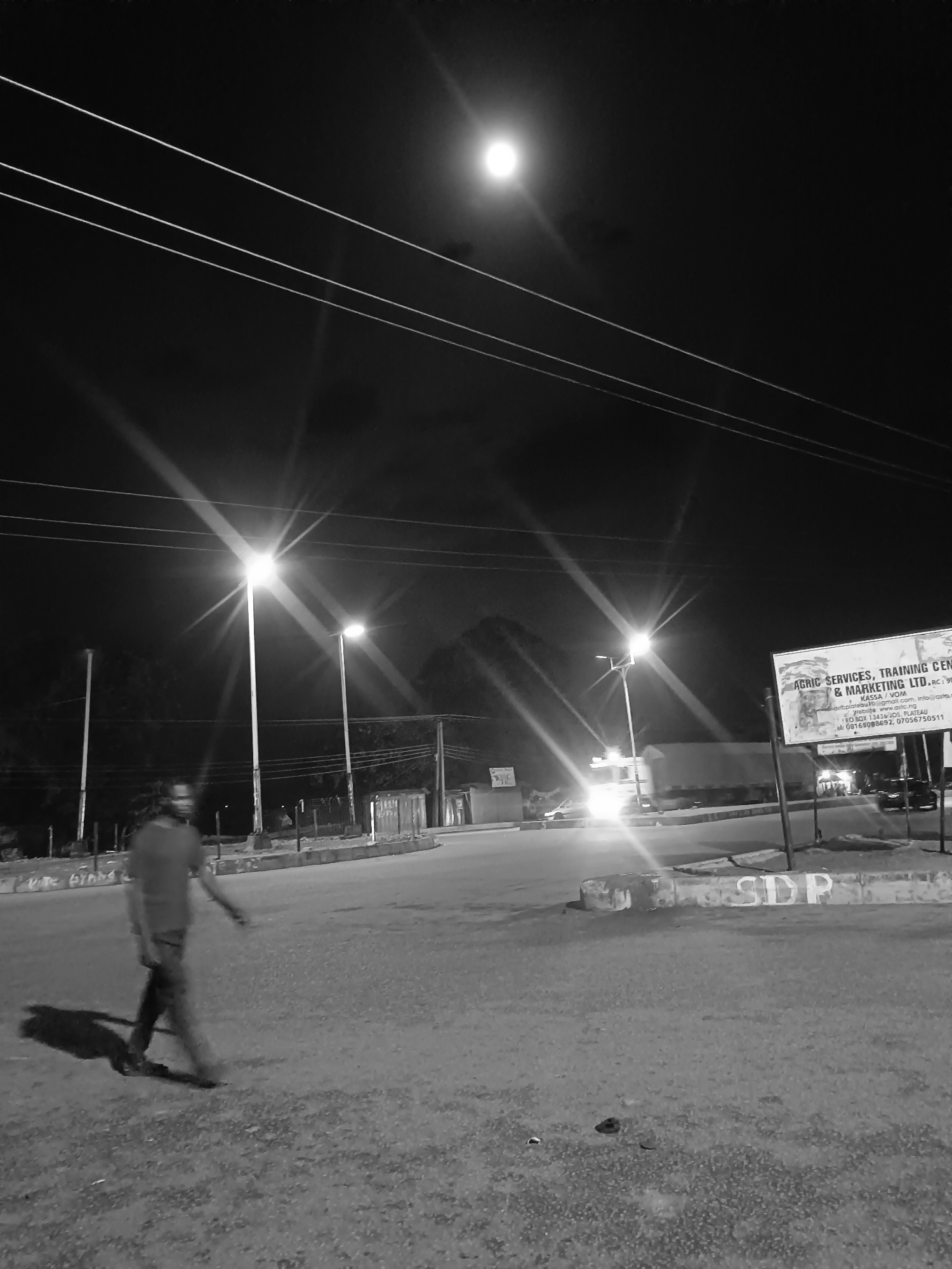 The image shows a night scene in Bukuru, Nigeria, illuminated by streetlights and the moon. A man is walking across an intersection. There are several streetlights, power lines overhead, a billboard for Agric Services, Training Center, and Marketing LTD, and a vehicle with its headlights on in the background. "SDP" is painted on the curb. The scene is desaturated, appearing in grayscale.