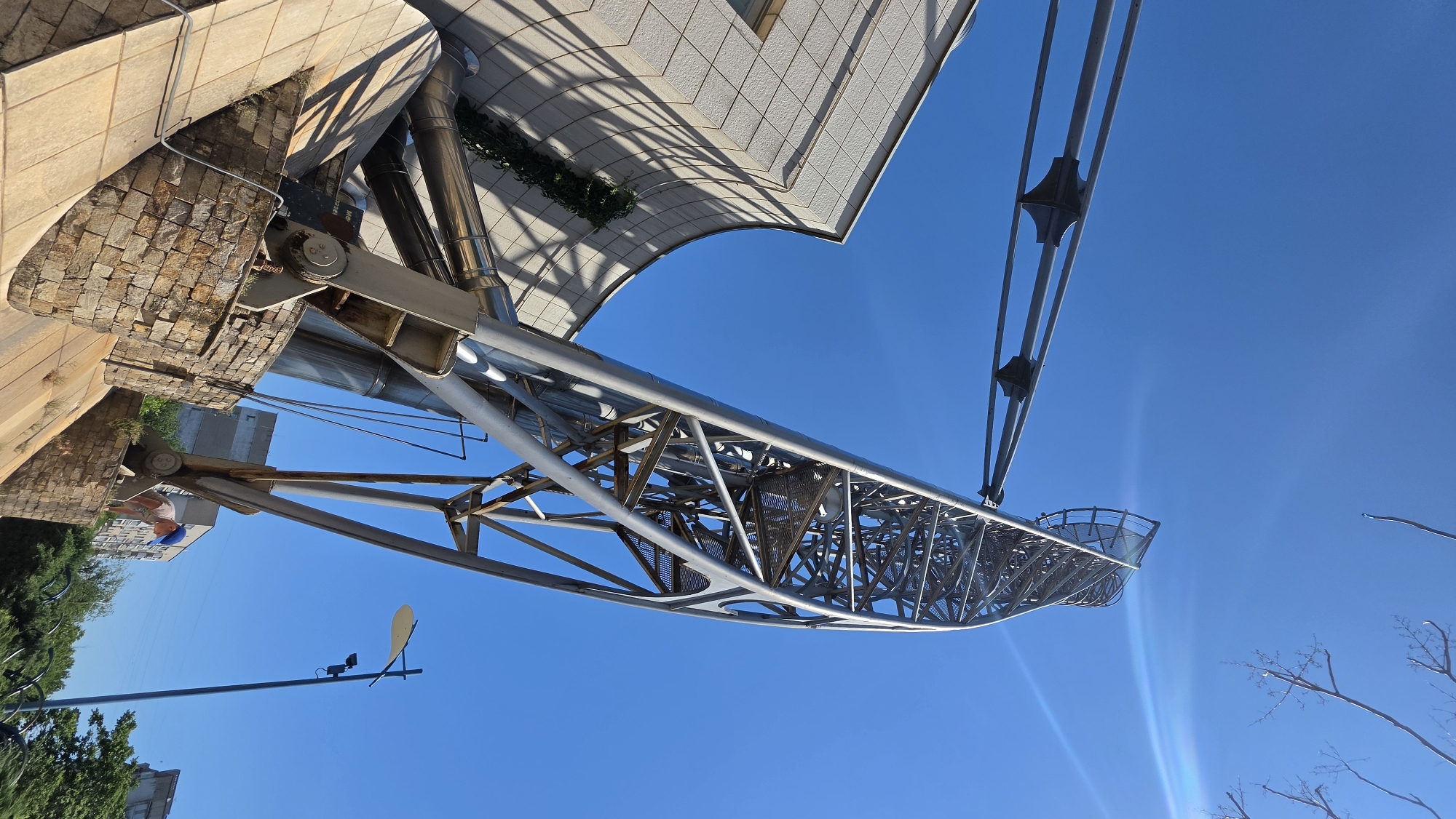 An upward-angle, full shot captures a modern, metallic sculpture on a sunny day in an urban environment. The sculpture, a tall, twisted structure with a helical design, dominates the right side of the frame, reaching towards a clear blue sky. Its metallic framework is intricate, with visible beams and supports forming a complex lattice. A curved, beige-tiled building with visible windows and shadows cast by its facade occupies the left side of the image. The base of the sculpture is anchored on a tiered structure made of rough, light-colored stones and concrete. A person wearing a blue cap is partially visible on the lower right, interacting with the stone base. In the background, beyond the sculpture and building, other urban buildings and lush green trees are visible under the bright sky. A street lamp with a curved fixture and a surveillance camera stands to the right of the sculpture. A thin branch with no leaves extends from the top right corner of the frame.