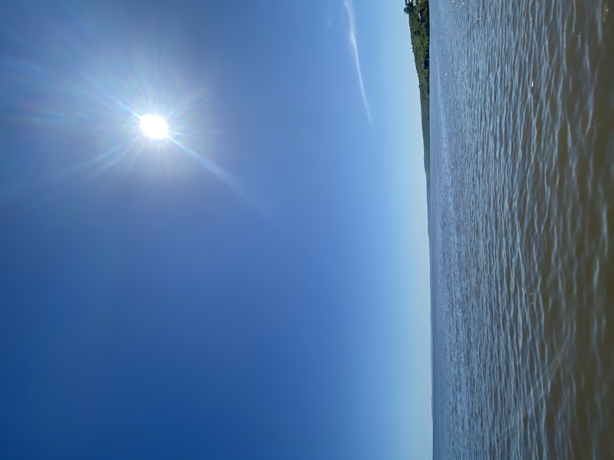 The image depicts a sunny day at the Volga River in Ulyanovsk, Russia. The sky is a clear, gradient blue, with the sun shining brightly in the upper left portion of the frame. To the right of the sky is a view of the Volga River's water, showing slight ripples and a dark-grey-blue hue. In the distance on the river's edge, there is a narrow line of green vegetation, possibly trees, indicating the shoreline. There are no visible people or other objects in the scene.