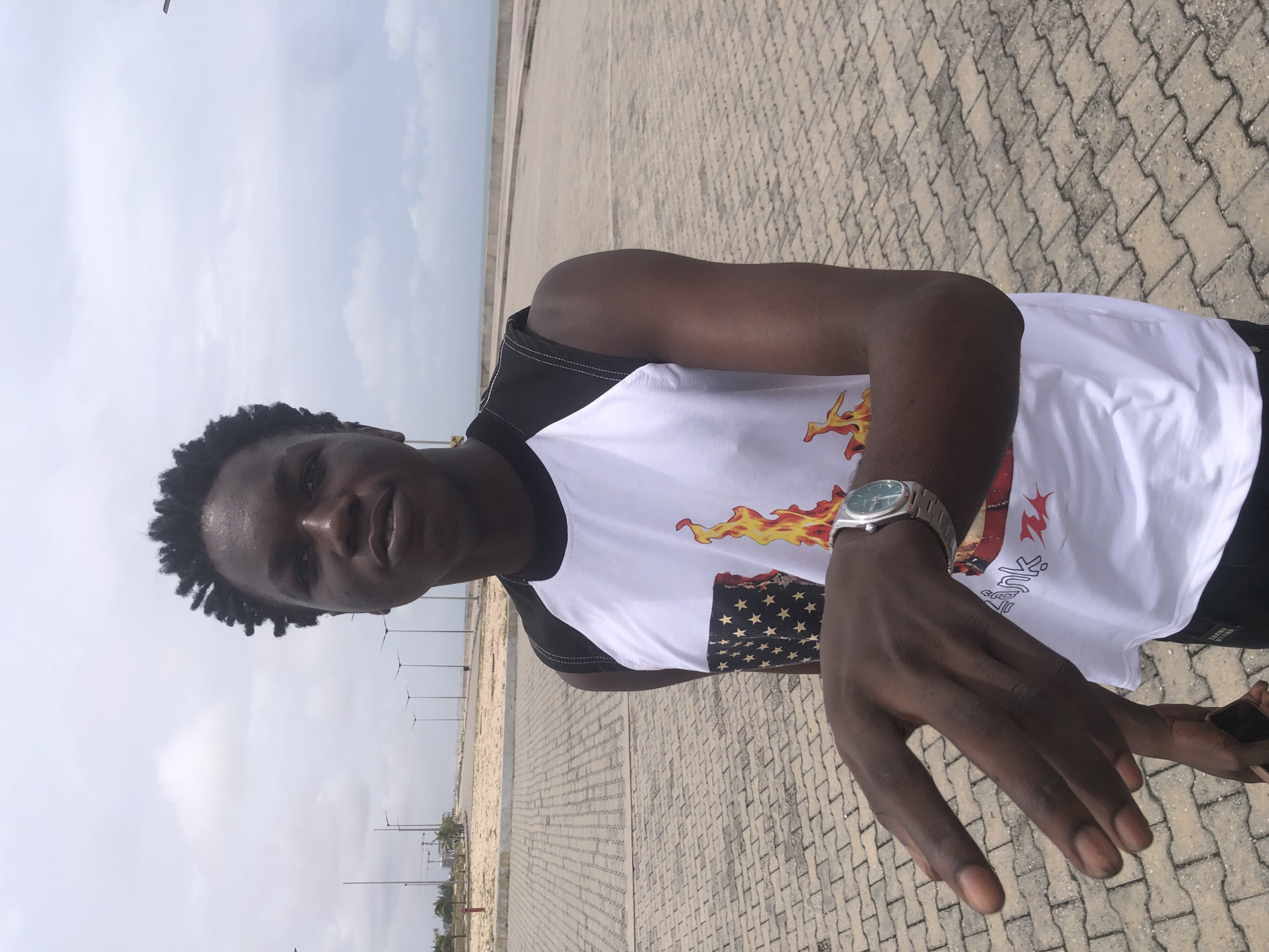 A young Black man stands outdoors in Ikoyi, Nigeria. He has dark skin, short, dark, curly hair, and is wearing a white sleeveless shirt with black trim. The shirt has a design featuring flames and an American flag. He is also wearing a silver watch on his left wrist. His left hand is extended towards the camera, with his fingers slightly curled. He is standing on a paved area made of interlocking bricks. In the background, a paved walkway runs parallel to a body of water, with a concrete barrier separating the two. Further in the distance, there are streetlights and some buildings under construction. The sky is overcast with scattered clouds.