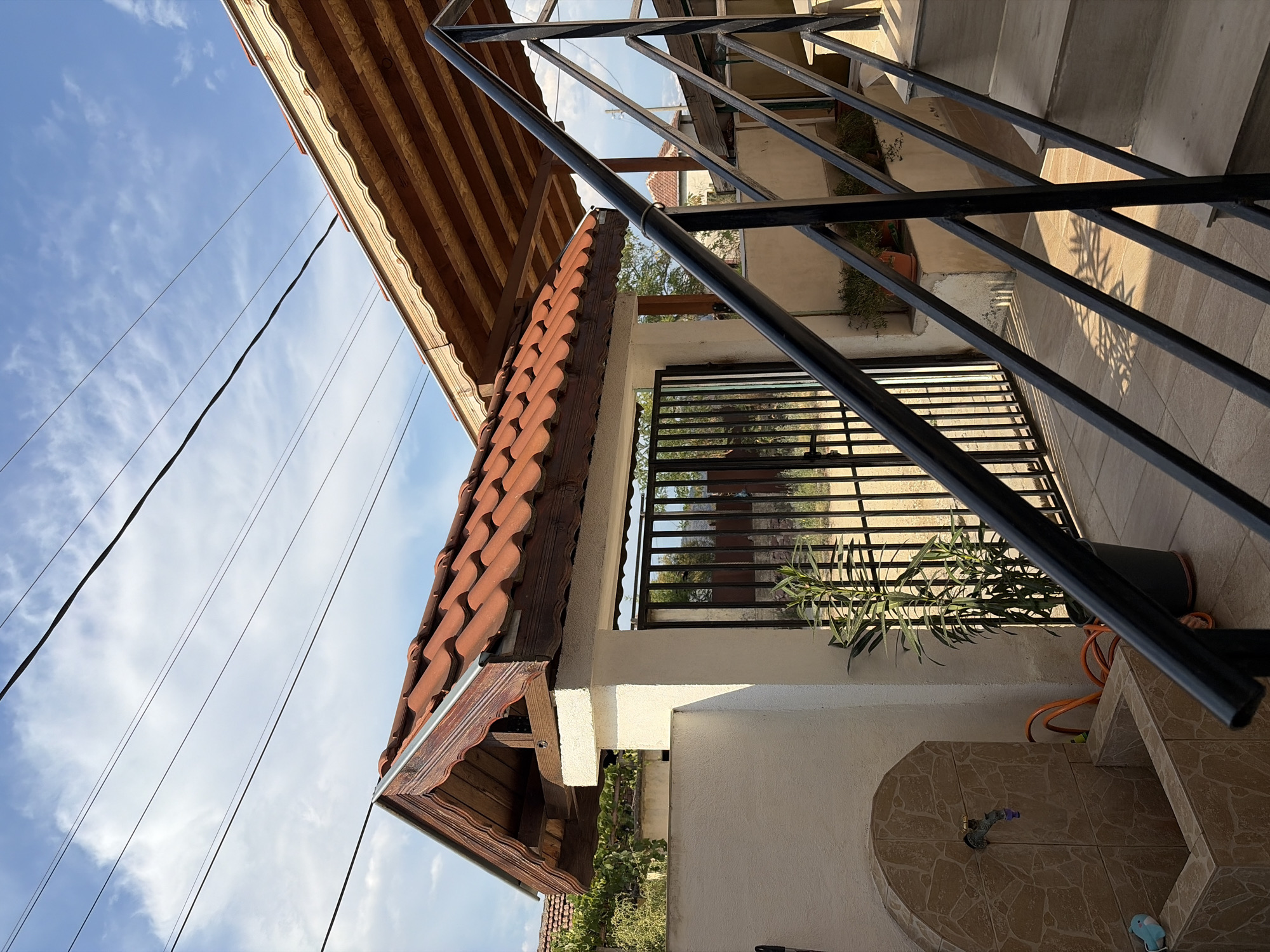 The image shows an exterior view of a property, likely a residential dwelling, under a bright blue sky with scattered white clouds. Overhead, several power lines are visible. In the foreground, a black metal railing runs diagonally up a set of outdoor stairs. To the left of the stairs is a white wall supporting a roof structure covered with terracotta tiles, featuring decorative wooden trim underneath. Adjacent to this is a black metal gate, behind which is a grassy area with some vegetation and trees. A tall potted plant stands to the left of the gate. Further to the left, a tiled water feature with a faucet is mounted on the white wall, with an orange hose coiled nearby. The ground is paved with light-colored tiles. The overall impression is that of a sunlit, outdoor space in a seemingly rural or suburban setting, possibly in Mitino, Bulgaria, given the context provided. There are no people or animals depicted in the image, and no discernable actions or interactions are occurring.