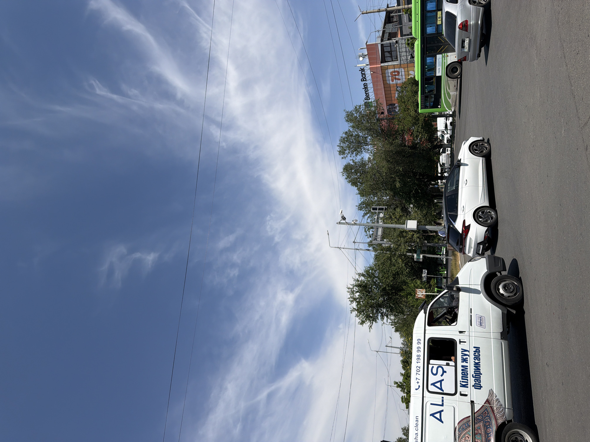 In Shymkent, Kazakhstan, a cloudy day features a street scene with several vehicles. A white van with "AL.AS" and Cyrillic text is on the left, with a graphic of a rug on its side. Next to it, a white car with a black roof is turning. Further down the road, a green bus with a white stripe and a "70" logo is visible. A silver sedan is in the right lane. A building with "Bereke Bank" branding is in the background, and traffic lights and trees are also present. The sky is a pale blue with wispy white clouds.