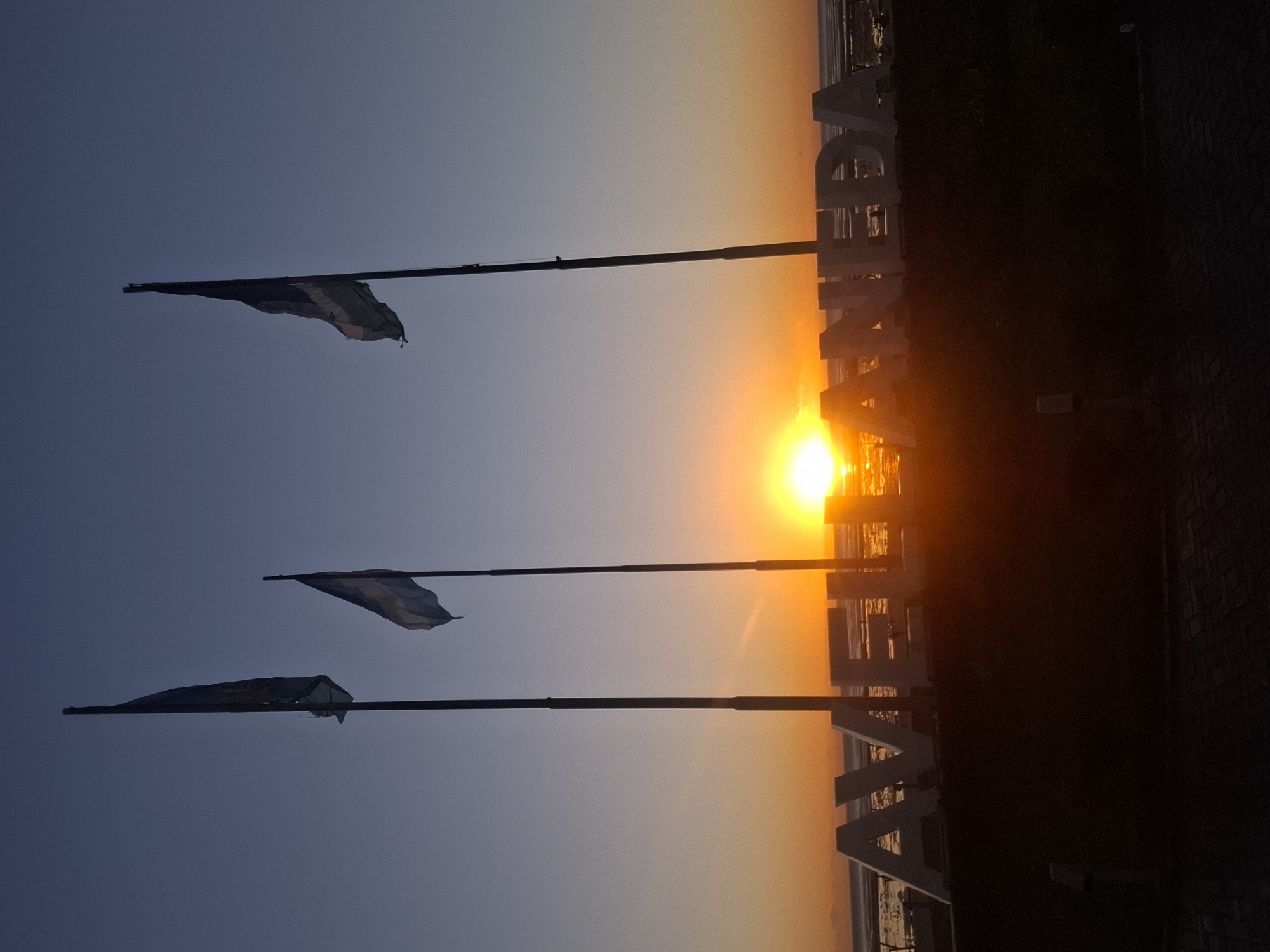 The image is a horizontal view of a sunset scene in Villa Domínico, Argentina. Three flagpoles stand in the foreground, each bearing a flag. The sky transitions from a dark blue to a golden orange as the sun sets on the horizon. In the foreground, the word "AVENIDA" is written in large, white letters. The sun is obscured by the letters, creating a silhouette effect. The scene is bordered by dark pavement on the right side. The sea is visible beyond the letters.