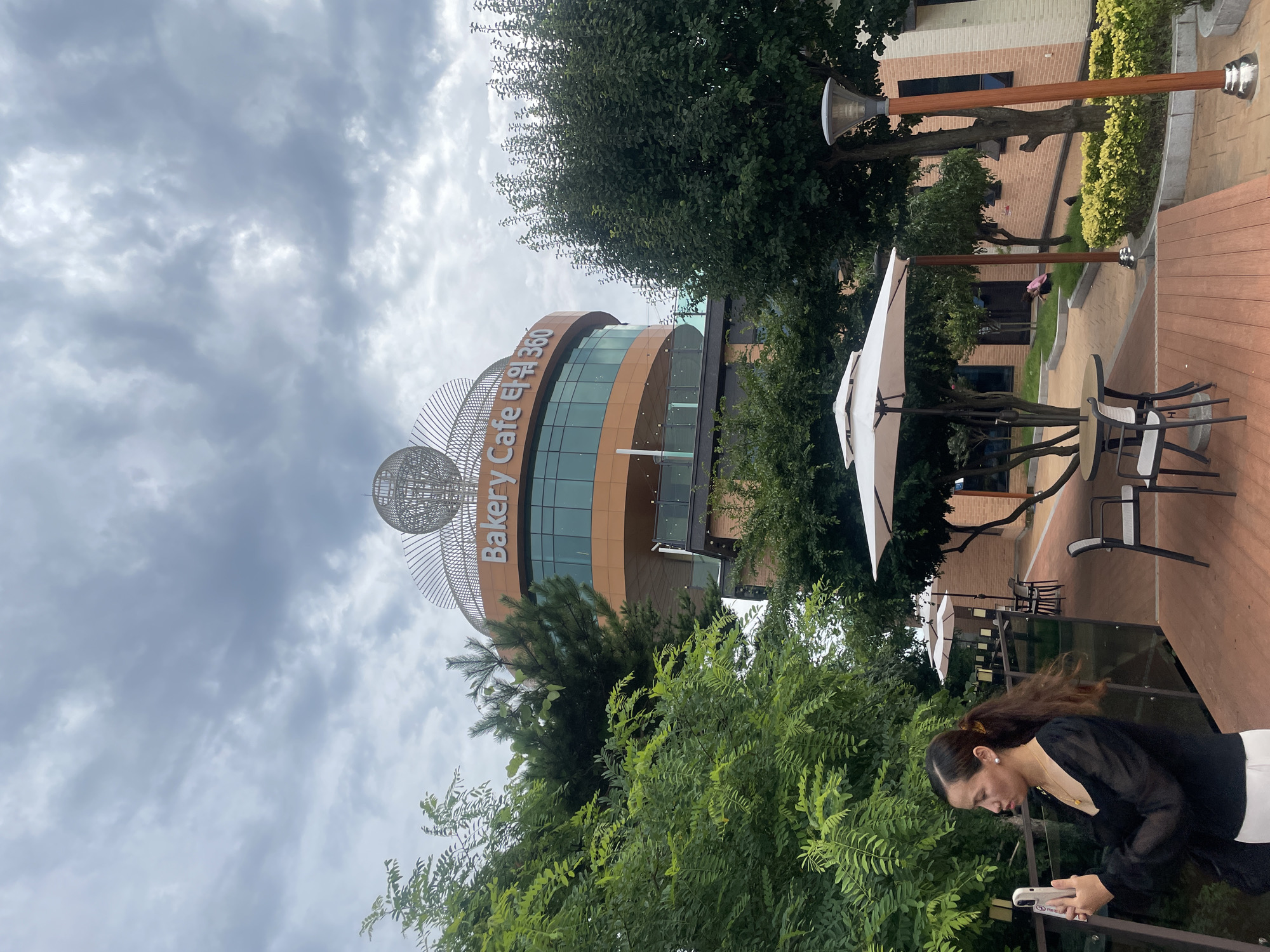 The image shows the exterior of a bakery cafe in Ansan, South Korea. The cafe is a multi-story building with a distinctive curved design and a large spherical ornament on its roof. The words "Bakery Cafe 타워 360" are visible on the facade. In the foreground, a woman in a black top and white pants is holding a smartphone, appearing to take a picture. She has her hair tied back in a ponytail with a yellow elastic band. Outdoor seating areas with white umbrellas and tables are visible, along with trees and greenery. The sky is overcast with dramatic clouds. The ground is paved with reddish-brown wooden planks in the foreground and patterned tiles further back. A lamppost with a modern design stands on the right side of the image. The overall scene suggests an outdoor seating area for a cafe or restaurant.