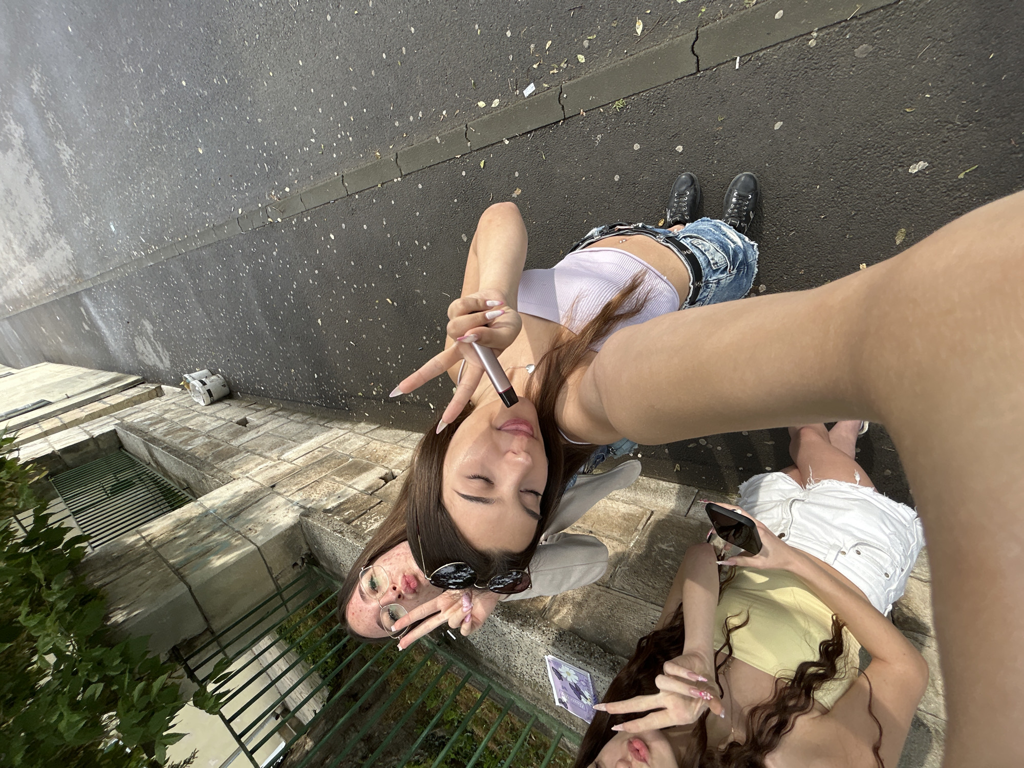 This media file is a selfie taken by two young women outdoors on a paved surface with a curb and some greenery. The woman closer to the camera is wearing a light purple tank top, distressed denim shorts, and black boots. She is holding a silver object up to her face and making a peace sign with her fingers. She has long brown hair and is wearing sunglasses. The second woman is on the left side of the frame, wearing glasses, a light yellow top, and white shorts. She is also making a peace sign with her fingers. The scene appears to be a street or sidewalk. The lighting suggests it is daytime, possibly sunny, as there are distinct shadows. The overall mood seems casual and friendly, given the selfie and the hand gestures. There is no visible text in the image.