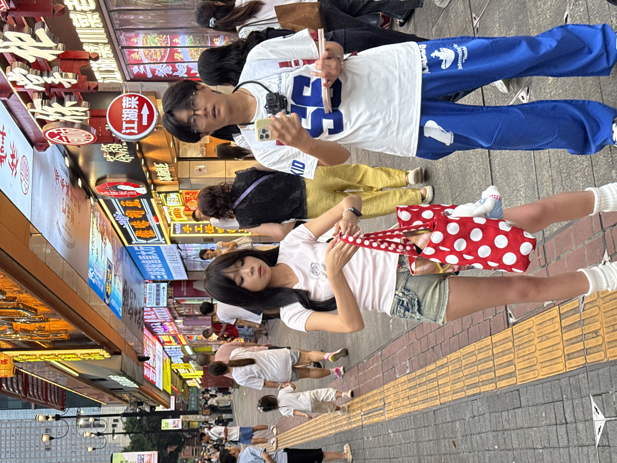 In Yuzhong District, China, on a bustling pedestrian street lined with brightly lit shops, a young man wearing glasses and a white t-shirt with a blue number "56" emblazoned on it, holds a smartphone and a small stack of pink papers. Beside him, a young woman with long dark hair wears a pink t-shirt and denim shorts, carrying a red polka-dot bag. Several other individuals, including children, are also visible walking along the street, which features a yellow tactile paving system alongside brickwork. The background is dominated by multi-story buildings adorned with colorful signs and advertisements.
