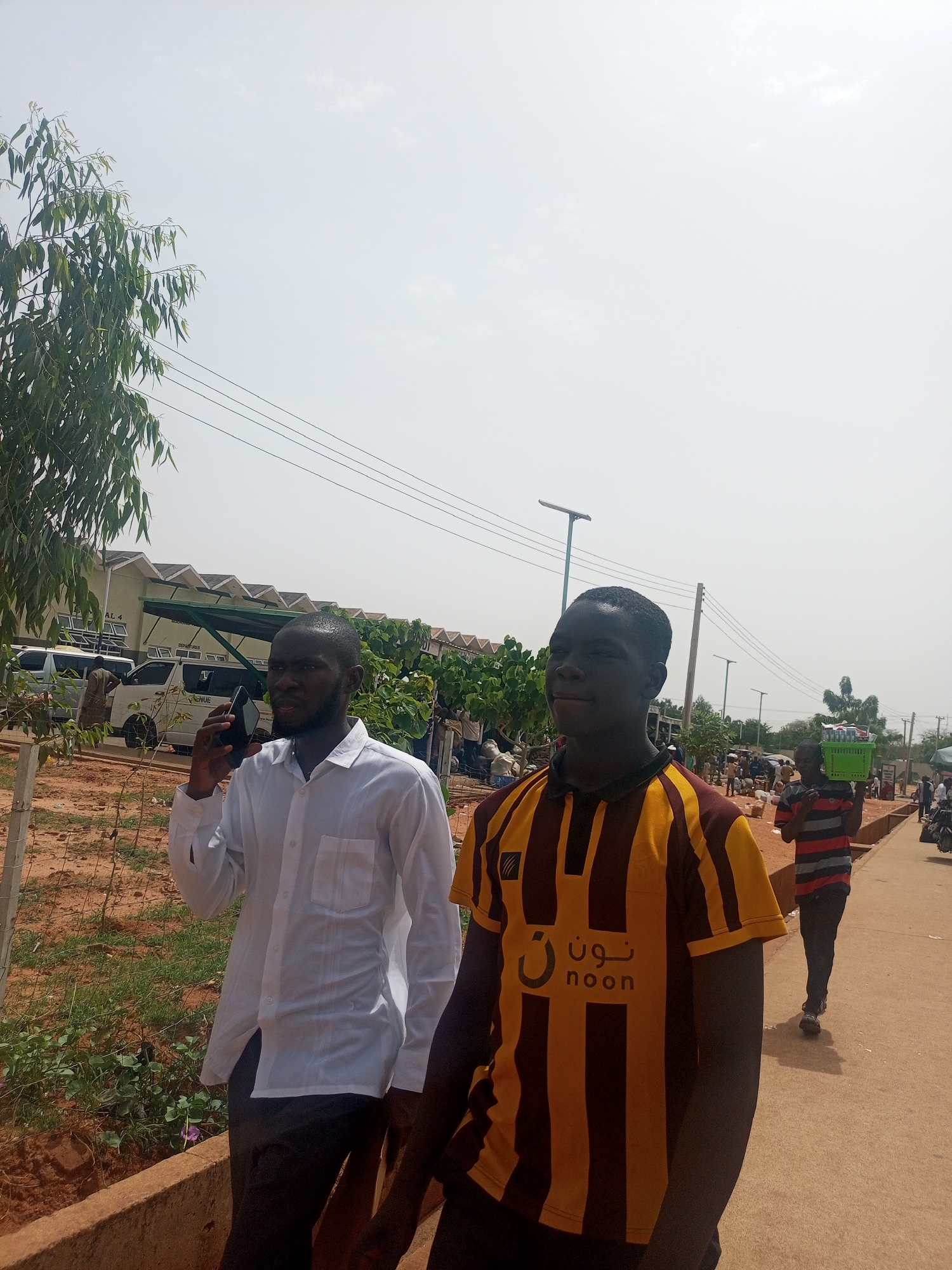 This outdoor image, taken in Gombe, Nigeria, features two men prominently in the foreground on a bright, sunny day. The man on the left, wearing a white long-sleeved shirt and dark trousers, is holding a black smartphone to his ear, seemingly engaged in a call. He has a beard and is looking forward. Beside him, the second man wears a brown and yellow striped polo shirt, which displays the word "noon" in both English and Arabic script on the chest. He is looking slightly to his right.
The background reveals a bustling environment. Other people are visible further down a reddish-brown path, including one individual on the right carrying a green crate filled with what appears to be packaged goods on their shoulder. Utility poles, overhead wires, and streetlights line the path. To the left, a large building with a distinctive green roof structure and white vans parked nearby suggests an institutional or commercial complex. Faint text like "A.B.U." and "DEPARTMENT" can be seen on the building, and "VENUE" is visible on a van. Lush green foliage, possibly from a eucalyptus tree, frames the left side of the image, contrasting with the bright, almost white, sky. The ground consists of dirt and some sparse greenery, typical of an unpaved street or market area.