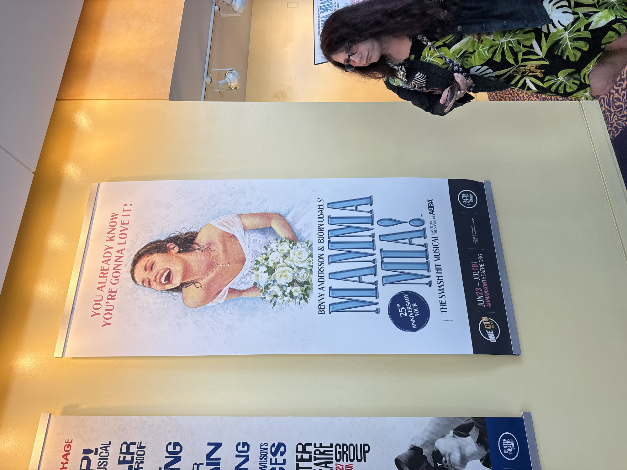 A woman with dark, curly hair and glasses stands to the right of a large poster advertising the musical "Mamma Mia!". The poster features a drawing of a laughing bride holding a bouquet of white flowers. The text on the poster reads "YOU ALREADY KNOW YOU'RE GONNA LOVE IT!", "BENNY ANDERSSON & BJÖRN ULVAEUS'", "MAMMA MIA!", "25TH ANNIVERSARY TOUR", and "THE SMASH HIT MUSICAL BASED ON THE SONGS OF ABBA". Below this, it shows "ONE CTG", "JUN23 - JUL19", and "AHMANSONTHEATRE.ORG". The poster is mounted on a pale yellow wall. To the left of the "Mamma Mia!" poster is another large poster with text that is partially visible. The woman is wearing a black jacket over a green and black patterned dress. She is holding a phone in her right hand. The environment appears to be indoors, possibly a theater lobby, with a patterned carpet visible on the floor.
