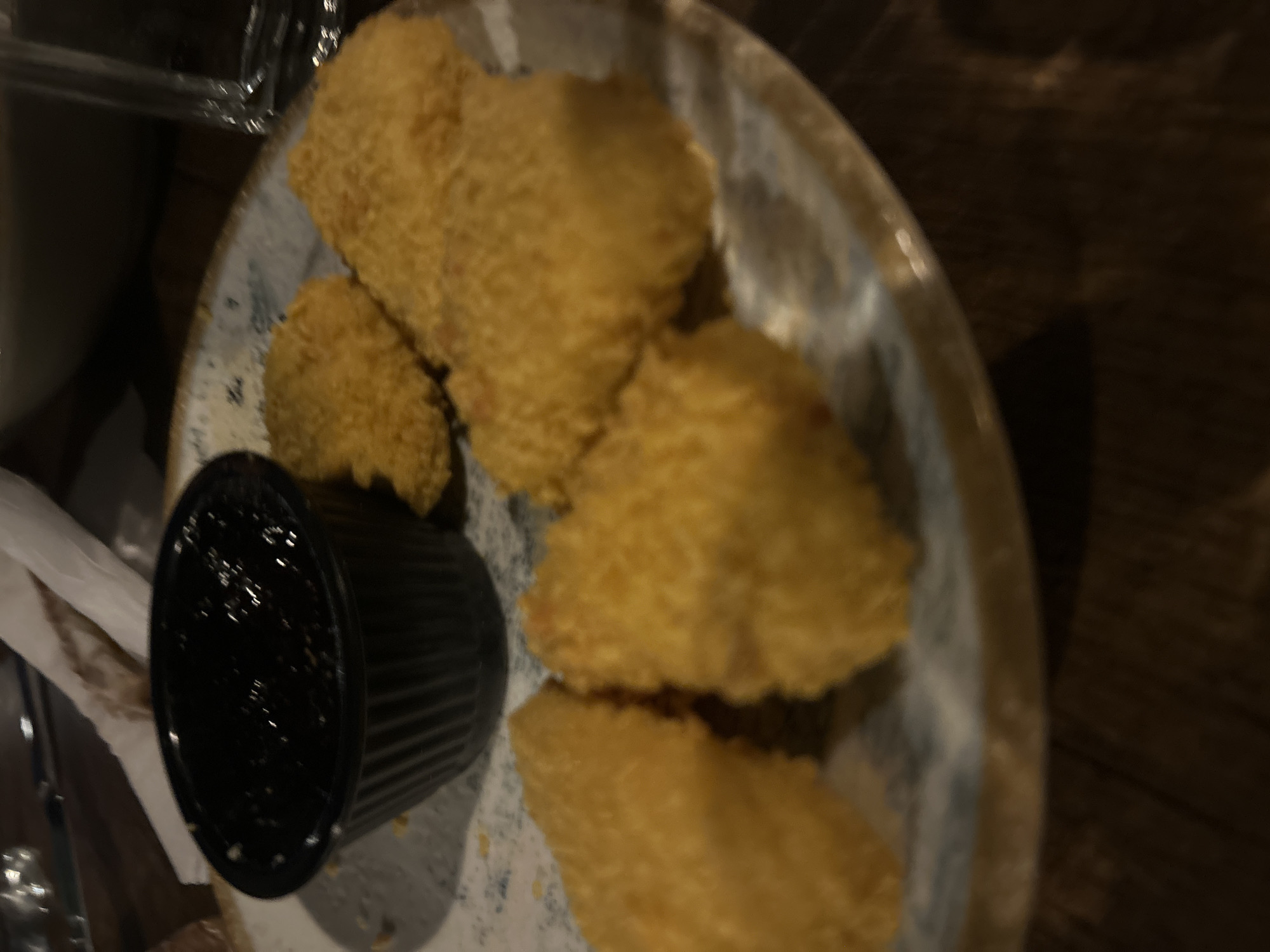 The image shows a plate of fried food items with a small black bowl of dark sauce on a table. The fried items appear to be golden brown and breaded, with a rough texture. They are arranged on a patterned plate. In the background, a glass of water and some napkins are visible. The lighting suggests an indoor setting, possibly a restaurant. The image does not provide any information about the city of Ruse, Bulgaria.