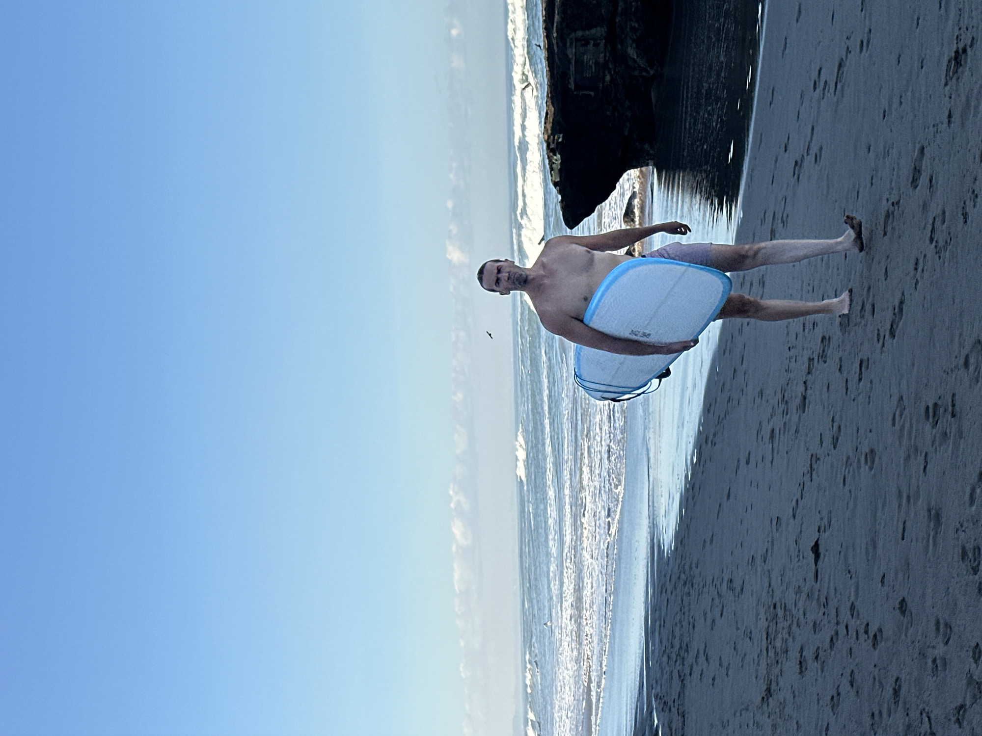A shirtless man with a surfboard walks on a sandy beach. The man is facing forward and looking at the camera. He is holding a white surfboard with blue trim. The beach is covered in footprints. In the background, the ocean has waves and there are birds flying in the sky. There is a large rock formation on the right side of the image. The sky is clear and blue. The image appears to be taken in the city of North Kuta, Indonesia.