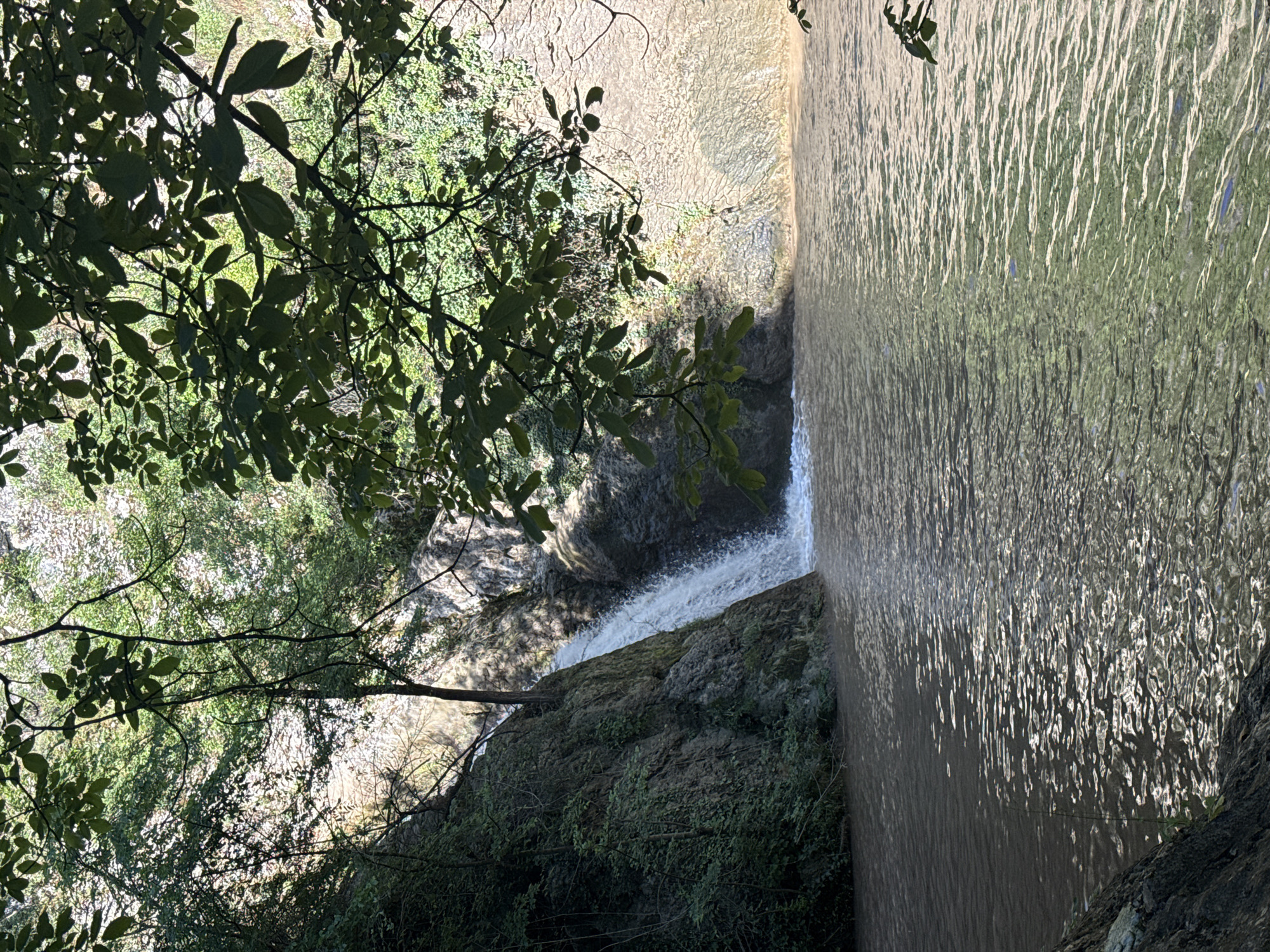 The image depicts a view of the Hotnitsa Waterfall in Hotnitsa, Bulgaria, from an elevated position. The waterfall cascades down a rocky cliff, partially obscured by trees and foliage. The water flows into a stream, reflecting light. Trees and vegetation surround the waterfall on both sides.