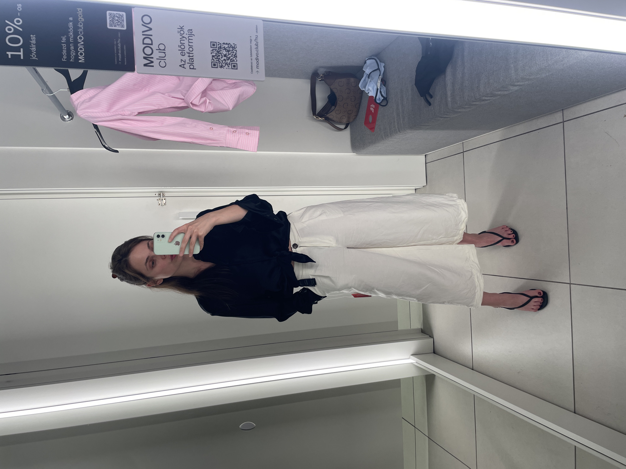 In a fitting room in Vecsés, Hungary, a woman stands in front of a mirror, taking a selfie with her phone. She is wearing a black knotted blouse and white wide-leg pants. On a clothes rack to her right, a pink pinstriped shirt is displayed. A grey bench sits against the wall, with a handbag, a red tag, and some folded clothes on it. The fitting room has bright lighting and tiled floors.