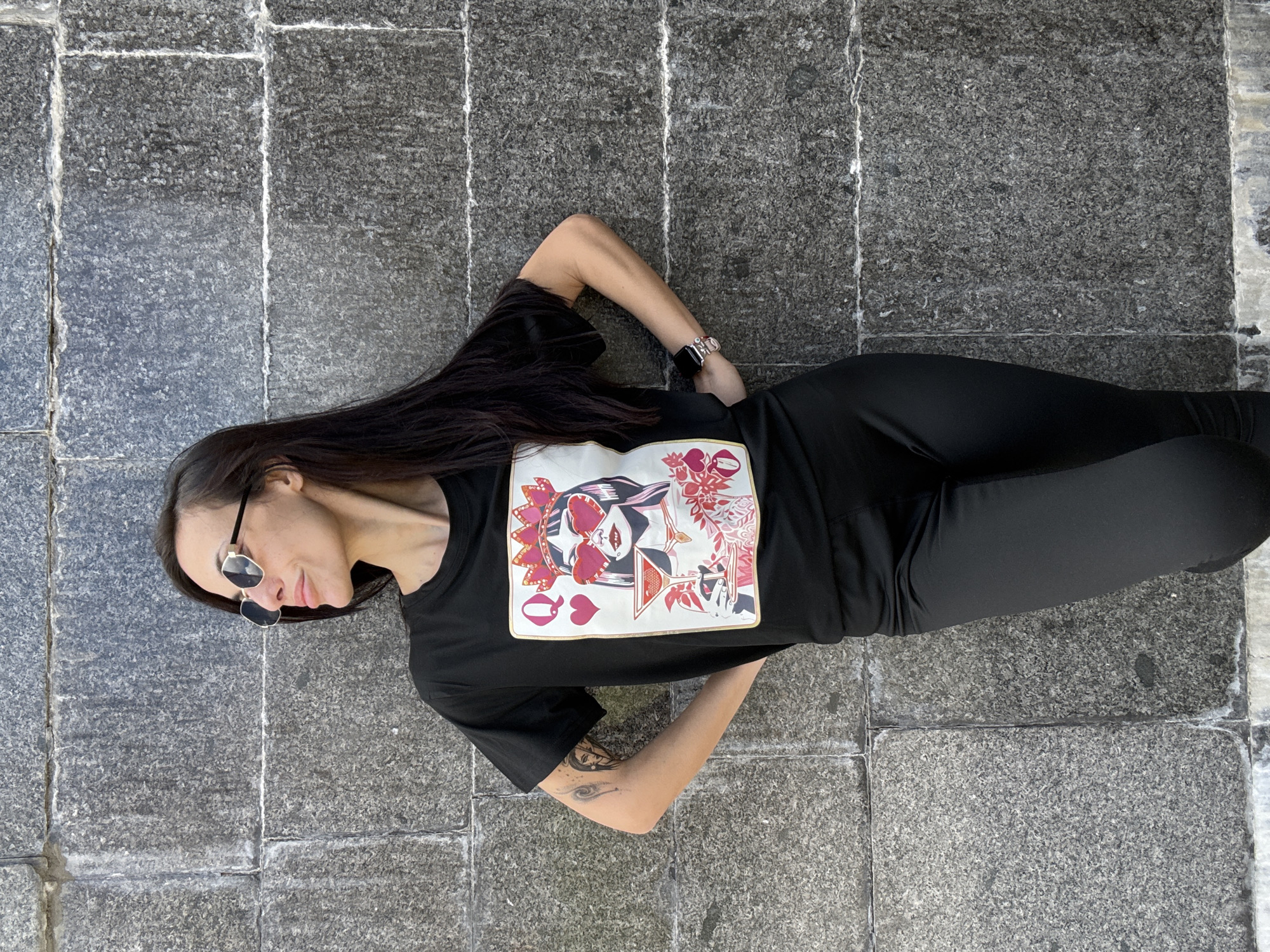 An adult female is depicted standing in front of a stone wall. She possesses long, dark hair and wears dark-framed, reflective sunglasses. Her attire consists of a black short-sleeved t-shirt featuring a stylized "Queen of Hearts" playing card graphic, which includes a figure with heart-shaped glasses, a crown, and a martini glass. She is also wearing black tight-fitting pants. Tattoos are visible on her right forearm, including an abstract design and a facial profile. On her left wrist, she wears a smartwatch with a metallic band. The woman is positioned with her body slightly angled to the left, her head turned further to the left, and her gaze directed horizontally to the left. Both hands are placed on her hips. The background is a wall composed of large, rectangular, dark grey granite blocks with a speckled texture. Lighter-colored mortar lines separate the blocks, exhibiting some weathering. The specific location within Pleven, Bulgaria, cannot be determined from the visual information provided.