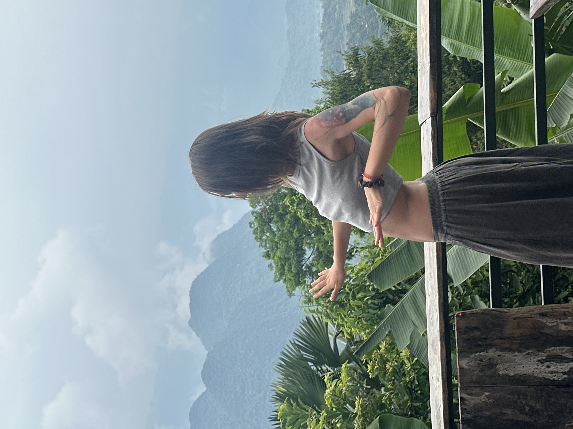 A woman is standing on a wooden balcony in Pù Luông Commune, Vietnam, with a scenic mountain and jungle view. She is wearing a gray tank top and dark gray pants. Her right arm is extended, and her left arm is bent, with her hand resting on her stomach. She has a tattoo on her right arm. The balcony is made of wood and overlooks lush green foliage and misty mountains. The sky is overcast with soft clouds. The overall scene suggests a serene and natural setting.