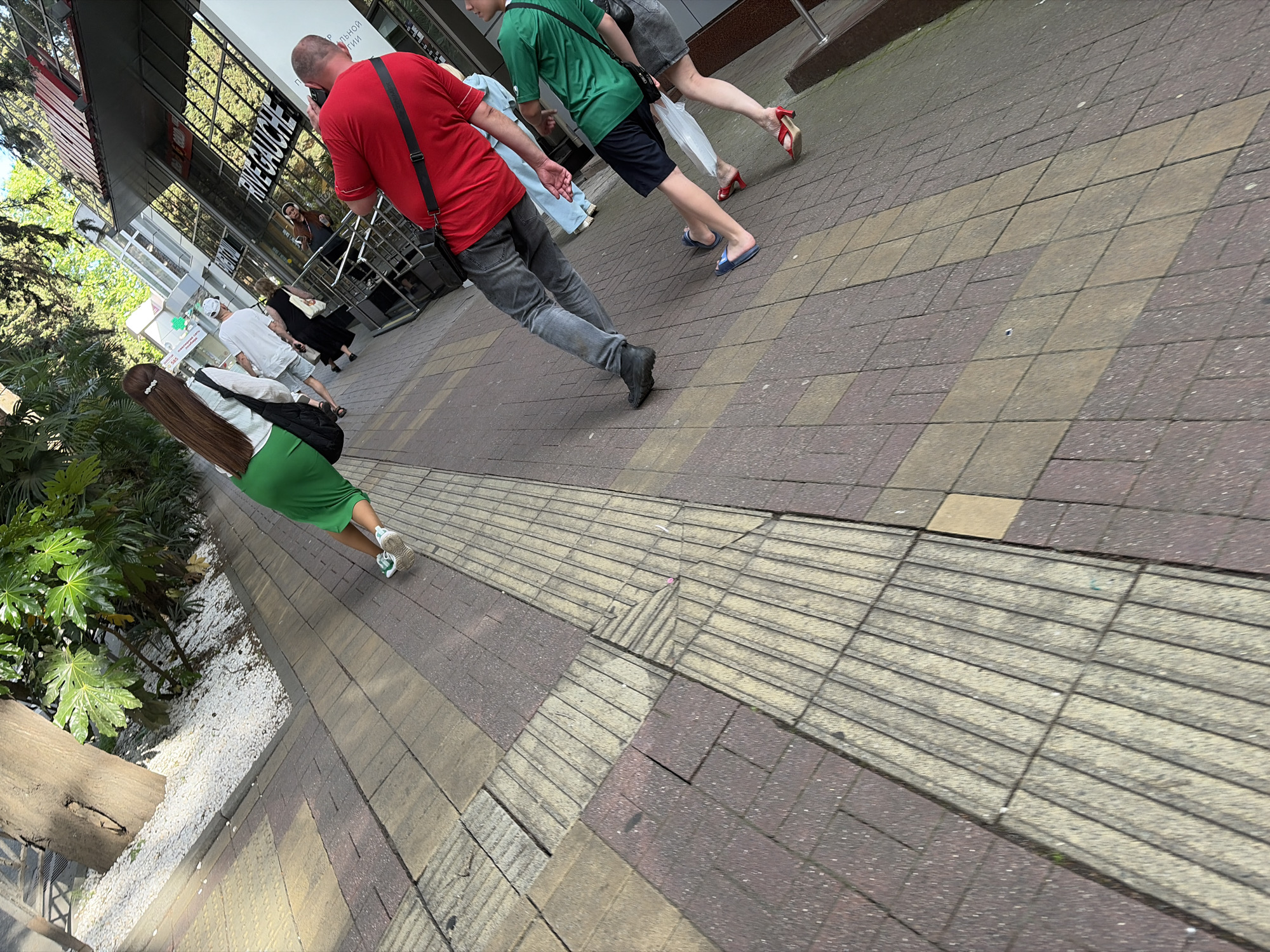 The image captures a dynamic street scene on a sunny day, likely in a city like Sochi, Russia. The photograph is taken from a slightly elevated, diagonal perspective, looking down a bustling paved sidewalk.
In the foreground on the left, lush green foliage and white pebbles line the edge of the sidewalk. A person with long brown hair, wearing a green skirt, white top, and white sneakers, walks away from the camera, carrying a black shoulder bag. The sidewalk itself is constructed from interlocking pavers in shades of reddish-brown and lighter beige, with distinct sections of tactile paving, featuring raised ridges, running diagonally across it.
Several people are walking along the sidewalk. In the mid-ground, a man in a red t-shirt, dark pants, and a black shoulder bag is visible, seemingly engrossed with his phone. Ahead of him, another person wearing a green t-shirt, dark shorts, and blue flip-flops also carries a black bag. Further down the path, a person in a grey skirt and red high-heeled sandals carries a white plastic bag. Other pedestrians are visible in the distance, near building entrances.
The background features a modern building with large, reflective glass windows. Clearly visible on its facade is the white text "РИВ ГОШ" (RIVE GAUCHE), indicating a cosmetics and perfumery store. Partially visible Russian text above it includes fragments like "ПАЛЬНОЙ" and "ТИИ," suggesting it might be part of a "Municipal Gallery." The bright daylight casts clear shadows, highlighting the active, urban environment.