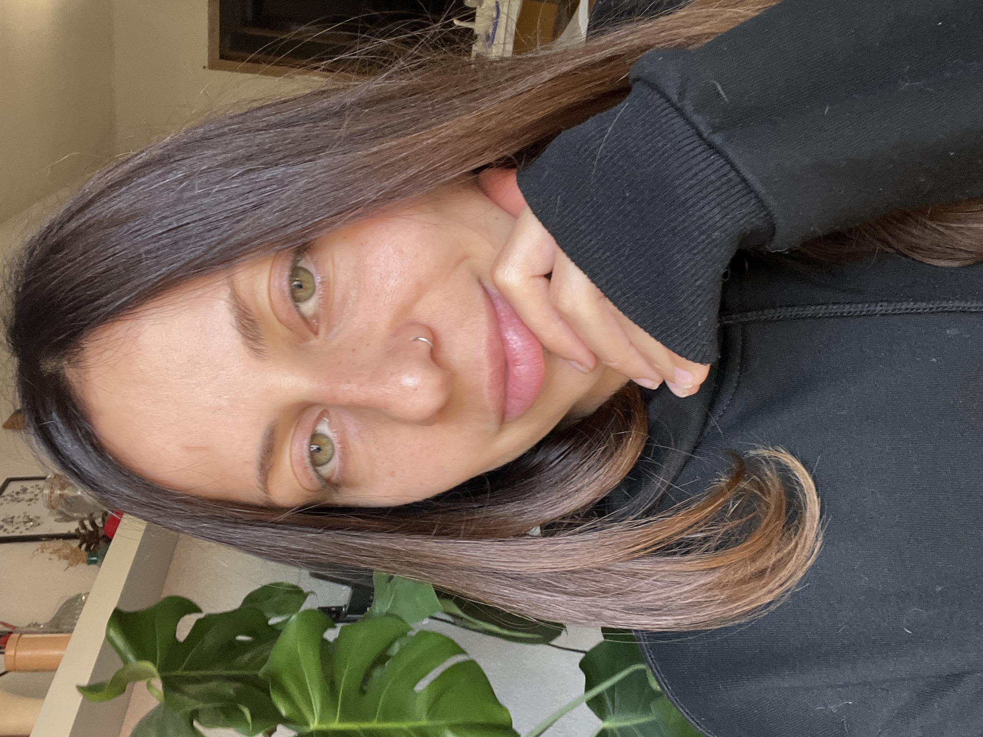 The image is a sideways close-up portrait of a young woman looking directly at the camera with a gentle, soft smile. Her long, dark brown hair, with subtle lighter brown or auburn highlights towards the ends, frames her face. She has fair skin with visible freckles and bright green eyes. A small silver hoop nose piercing adorns her left nostril. Her left hand is positioned delicately against her cheek and chin, with her arm partially covered by the cuff of a black hooded sweatshirt. The cuff and body of the sweatshirt show slight signs of lint.
In the foreground, a large, dark green leaf from a Monstera plant is prominently featured, suggesting an indoor setting. The background is softly lit and blurred, revealing a light-colored wall and indistinct furniture or decor, including what appears to be a pinecone on a shelf in the upper background. The overall atmosphere conveyed is calm and natural. The time of day appears to be daytime, with soft ambient light. No text is visible in the frame. The location context provided is Barcelona, Spain.