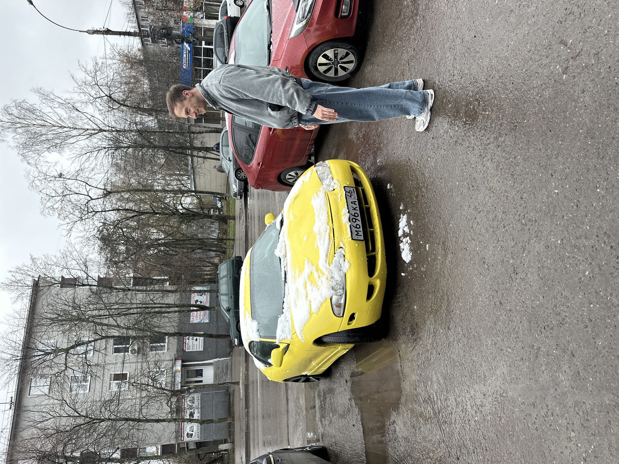 A man stands on wet pavement next to a yellow car covered with snow on its hood and windshield. The car has a license plate number M696KA 46. Another red car is parked behind it. The man is wearing a light grey jacket, jeans, and white sneakers. Bare trees line the street, and a grey building with several signs is visible in the background. The location is the city of Kursk, Russia.