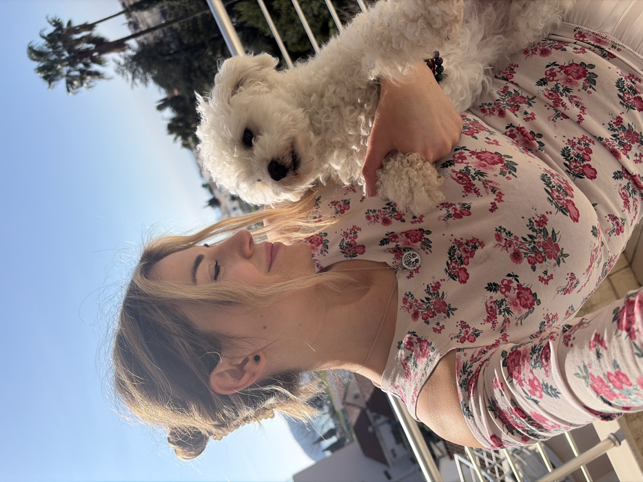An outdoor image depicts an adult female holding a small white curly-haired dog on a balcony in Budva, Montenegro. The female, with blonde hair styled in a bun and framing strands, wears a light-colored, long-sleeved top with a pink floral pattern and shoulder cut-outs. She has a silver necklace with a circular "Tree of Life" pendant, small dark stud earrings, and a dark beaded bracelet on her right wrist. Her head is angled to the right, eyes partially closed, with a subtle smile directed towards the dog held by her left arm against her chest. The dog faces forward. The background shows a clear blue sky, a metal balcony railing, buildings with red-tiled roofs, green foliage, and palm trees, with distant hills characteristic of a coastal town.