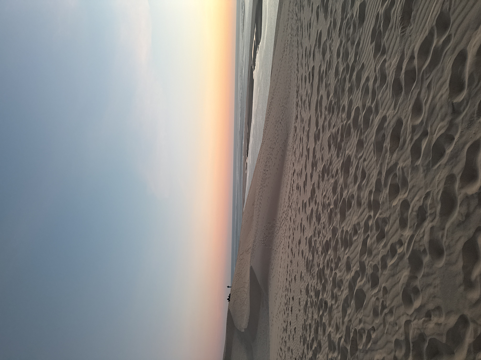 This is a landscape photograph of the Dune of Pilat in La Teste-de-Buch, France, during sunset. The foreground is dominated by rippled sand dunes covered in numerous footprints. In the midground, a line of people can be seen standing and sitting on the crest of a dune, silhouetted against the sky. Beyond the dunes, a body of water, likely the Atlantic Ocean, stretches to the horizon, where the sky transitions from a pale blue at the top to soft hues of pink and orange near the horizon, indicating the setting sun. The overall impression is one of a serene, natural environment at dusk.