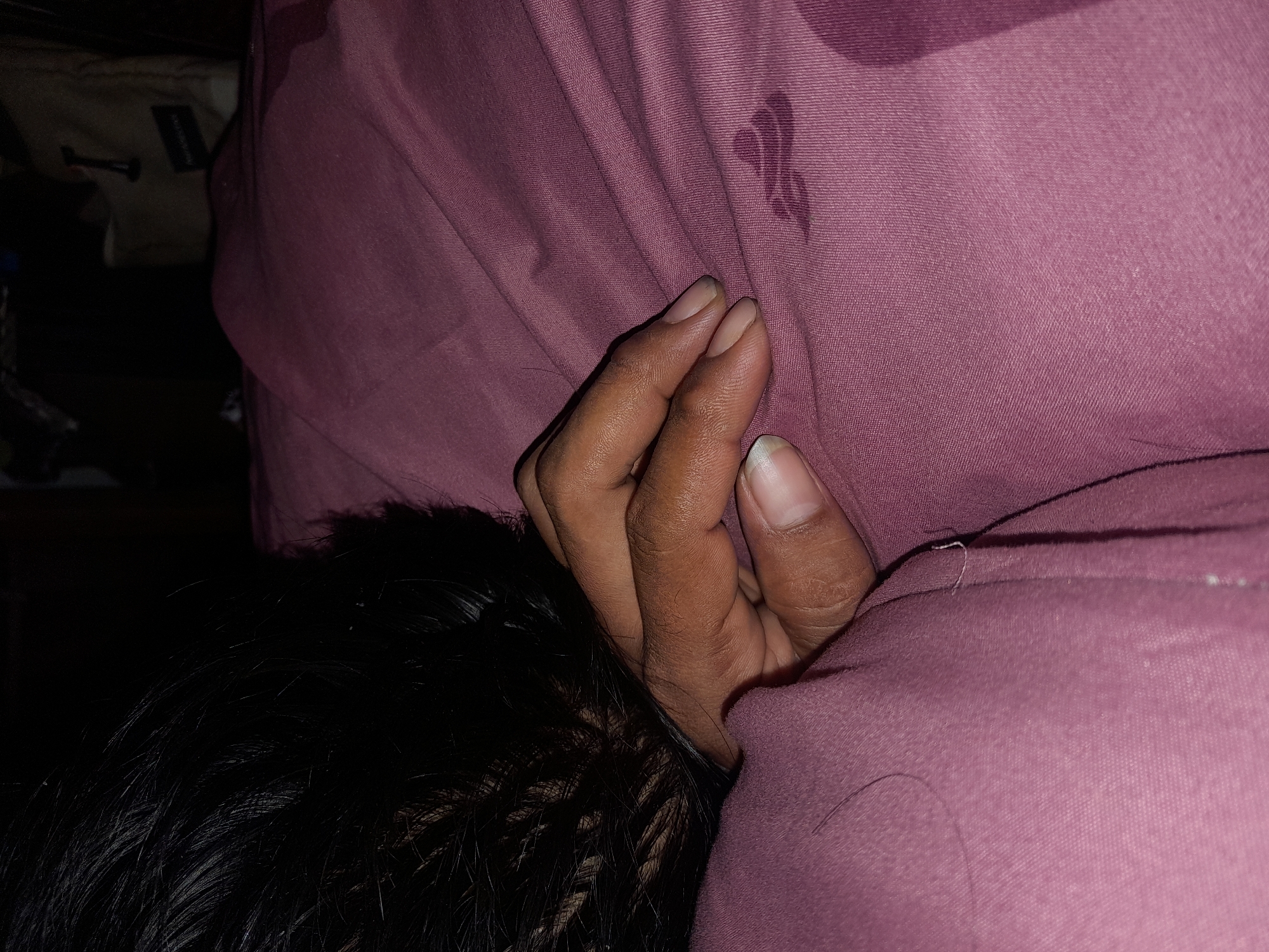 The image depicts a close-up view of a person's hand resting on a pink, textured fabric, likely bedding. The hand, with visible fingernails, is positioned on the fabric, which shows some wrinkling and a darker, possibly wet, stain. To the left, a portion of dark, possibly black hair is visible, obscuring part of the background. The overall lighting is dim, creating shadows and a sense of indoor illumination. The specific location in Keboharan, Indonesia, is not discernible from the visual information presented in the image.