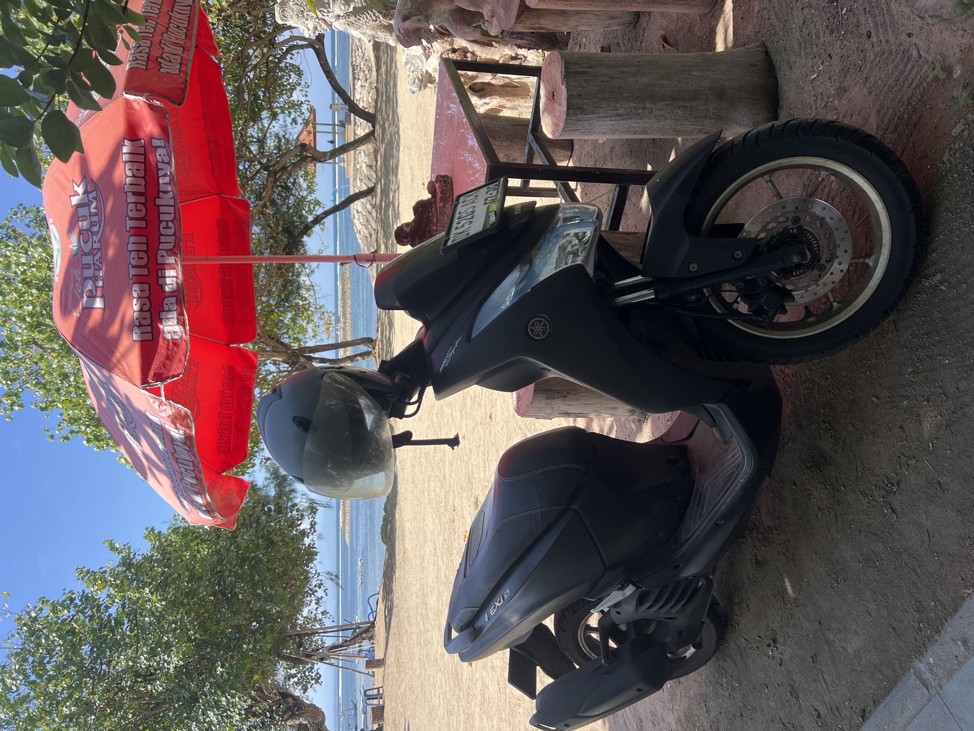A black motorcycle is parked on a sandy beach next to a red umbrella advertising "Pucuk Harum" tea. The umbrella is open and displays text in Indonesian that translates to "The best tea taste is at its peak!". Behind the motorcycle, a red table with metal legs is visible, supported by wooden logs. Further in the background, the calm blue ocean meets a sandy shore, with a few trees and beach chairs scattered around. The sky is a clear, bright blue, suggesting a sunny day. The scene appears to be a peaceful beachside setting in Denpasar, Indonesia, likely in the afternoon given the lighting. The motorcycle is a scooter-style model, with a helmet resting on its handlebars. The license plate is partially visible and reads "DK 3725 KCV".