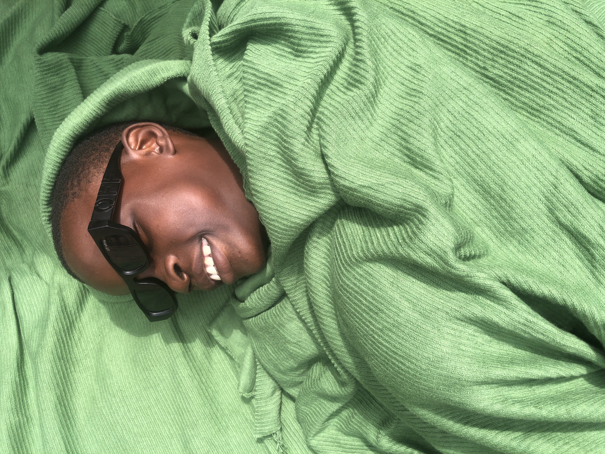 A close-up overhead shot shows a person lying down, wrapped in a green, ribbed fabric. The person is wearing black sunglasses with "OFF WHITE" written on them. The person's face is visible, with eyes closed and a smile on their lips, revealing white teeth. The green fabric appears to be a hooded garment or blanket, with folds and creases creating shadows. The lighting suggests an outdoor setting, possibly during the day. No specific location in Ado, Nigeria is discernible from the image itself.