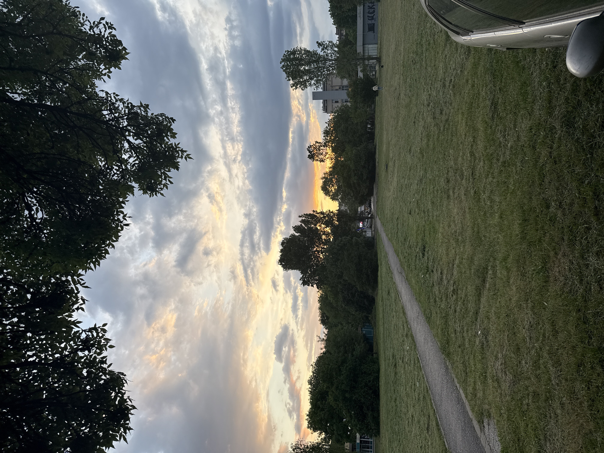 The photograph captures a scene at sunset in Sofia, Bulgaria. The sky is filled with clouds, tinged with yellow and orange hues near the horizon. The foreground consists of a grassy field with a narrow path running through it. A car is partially visible in the upper right corner. Trees border the field, with buildings visible in the background. The scene is peaceful and serene.