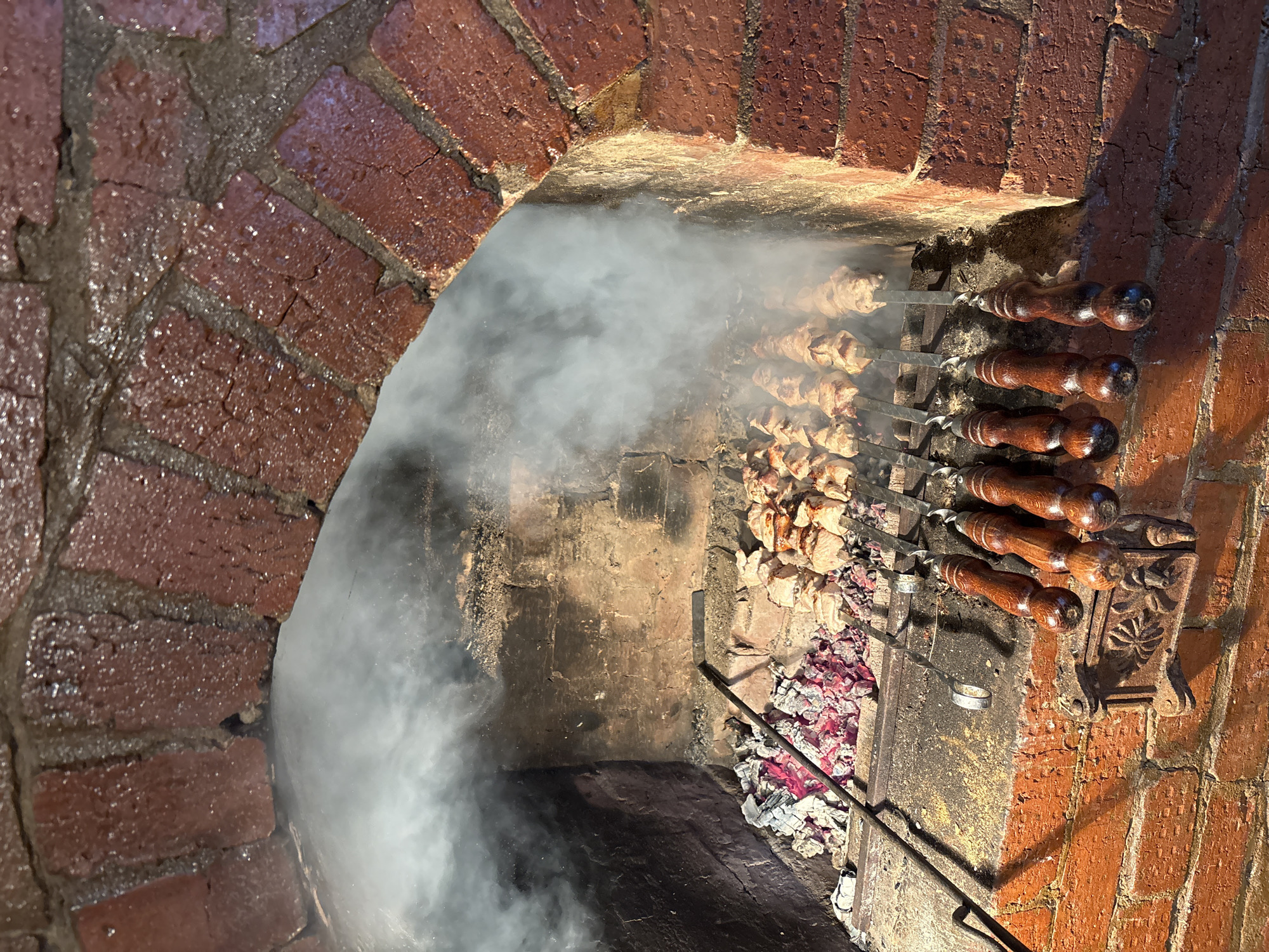 The image depicts an indoor brick oven in a dimly lit setting, filled with smoke. The oven is constructed of red bricks, with a visible arch opening. Inside, several skewers are arranged horizontally across the opening, laden with meat. Burning embers and partially burnt pieces of wood are visible at the bottom of the oven. The scene suggests the cooking of shish kebab, with the smoke obscuring much of the oven's interior. The location is confirmed as the village of Алегазово, Russia, based on geographical and cultural food indications.