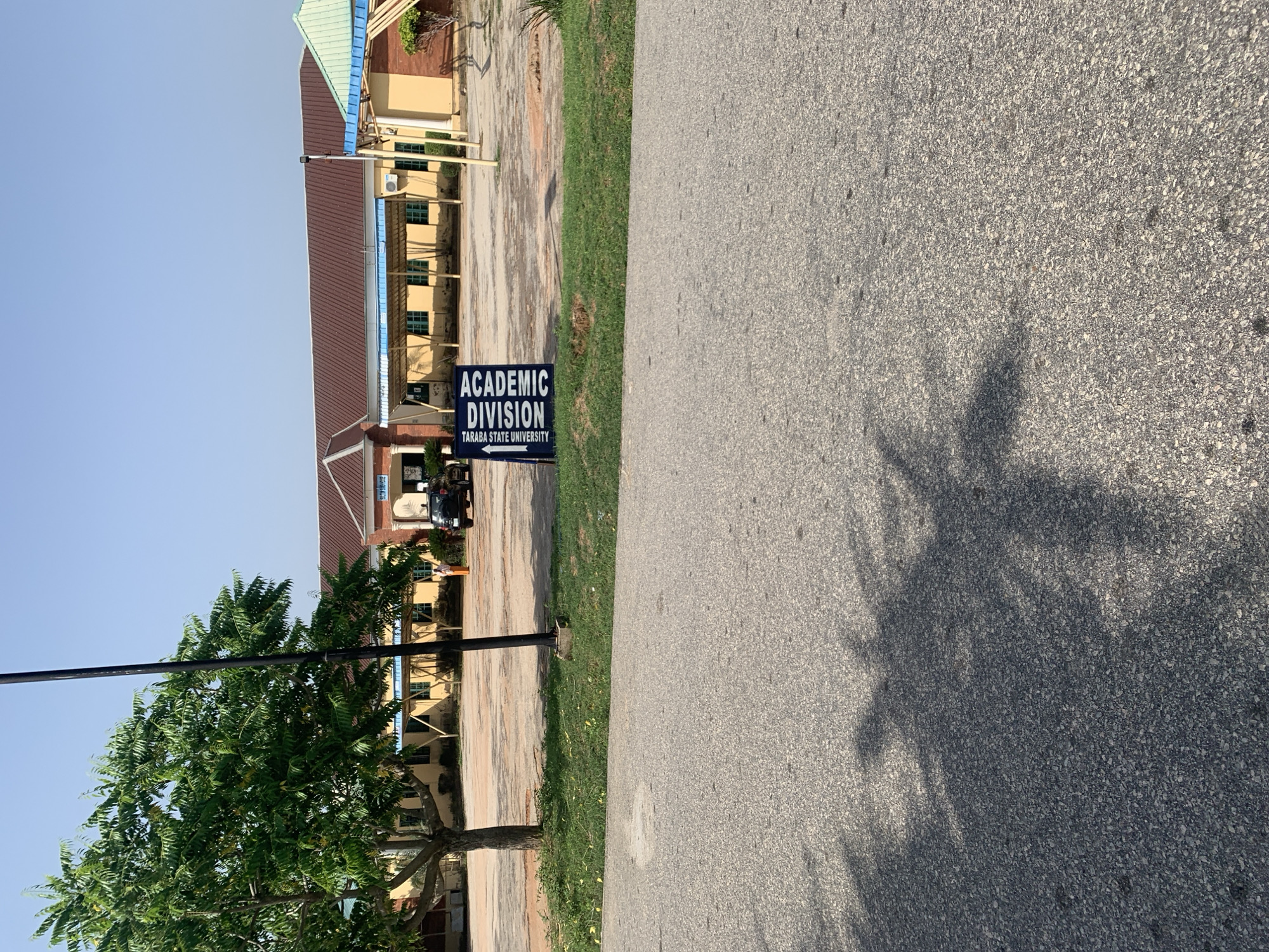 The image depicts an outdoor scene on a bright, clear day, likely in Jalingo, Nigeria, given the presence of a sign identifying "TARABA STATE UNIVERSITY." In the foreground, a paved road takes up the lower half of the frame, with tree shadows cast across it. To the left, a large, leafy green tree stands tall next to a black pole. In the middle ground, a light-colored building with a brown-tiled roof stretches horizontally across the image. In front of the building, a black SUV is parked. To the left of the SUV, a blue and white sign with black text reads "ACADEMIC DIVISION TARABA STATE UNIVERSITY." A person in an orange dress is visible standing near the building. The ground in front of the building is a mix of paved areas and dirt.