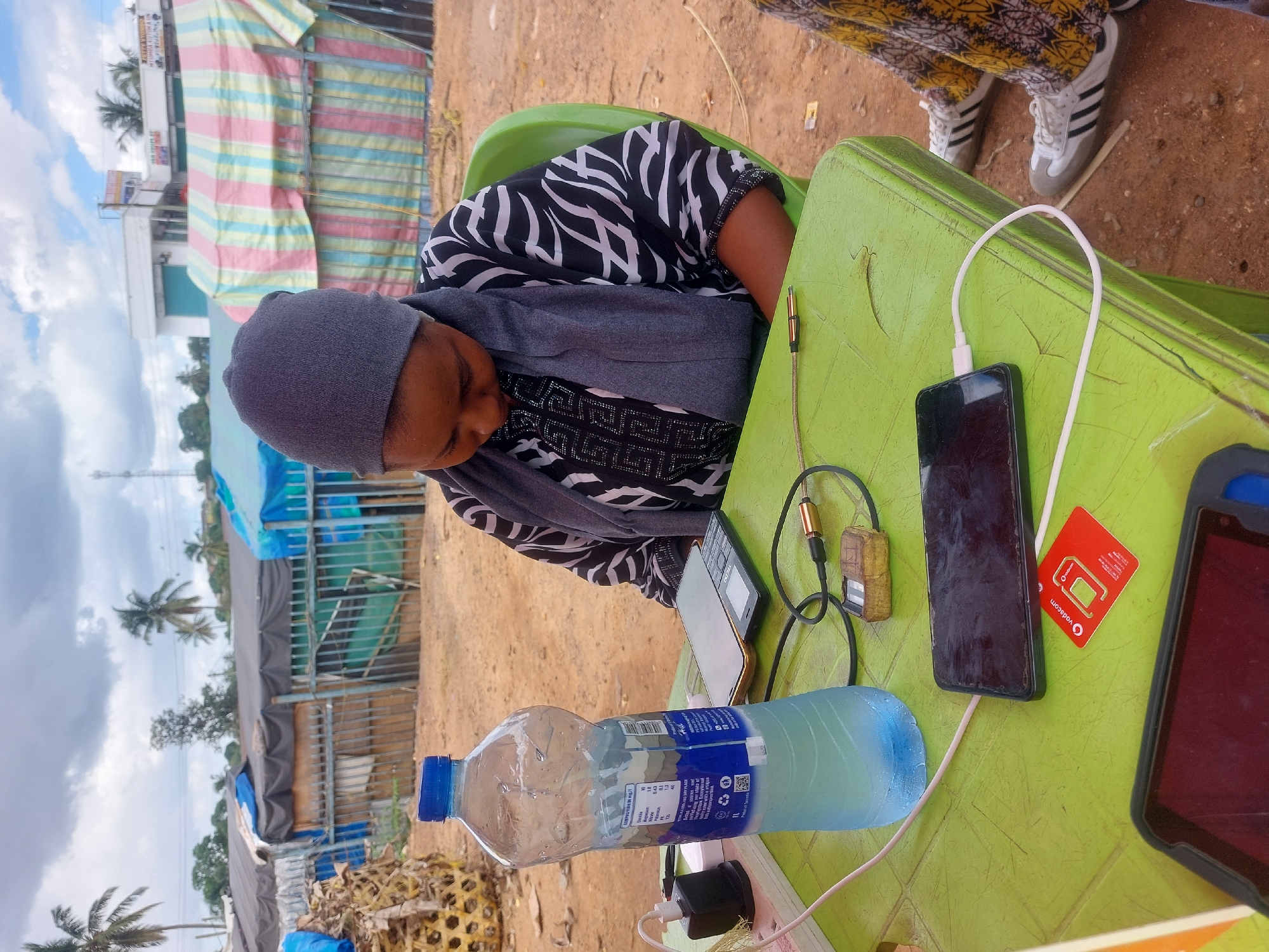 A woman wearing a grey head covering, a grey shawl, and a black and white patterned shirt sits at a light green plastic table. On the table, there's a mobile phone connected to a white charging cable, a red Vodacom SIM card, a bottle of water, a tablet in a black case, another phone, a connector, and a charging adapter. The table is positioned outside, with tents and buildings visible in the background, suggesting a possible market area. Considering the red Vodacom SIM card and the architecture in the background, the location is likely within Dar es-Salaam, Tanzania. Another person is partially visible in the upper right, wearing pants and Adidas sneakers.