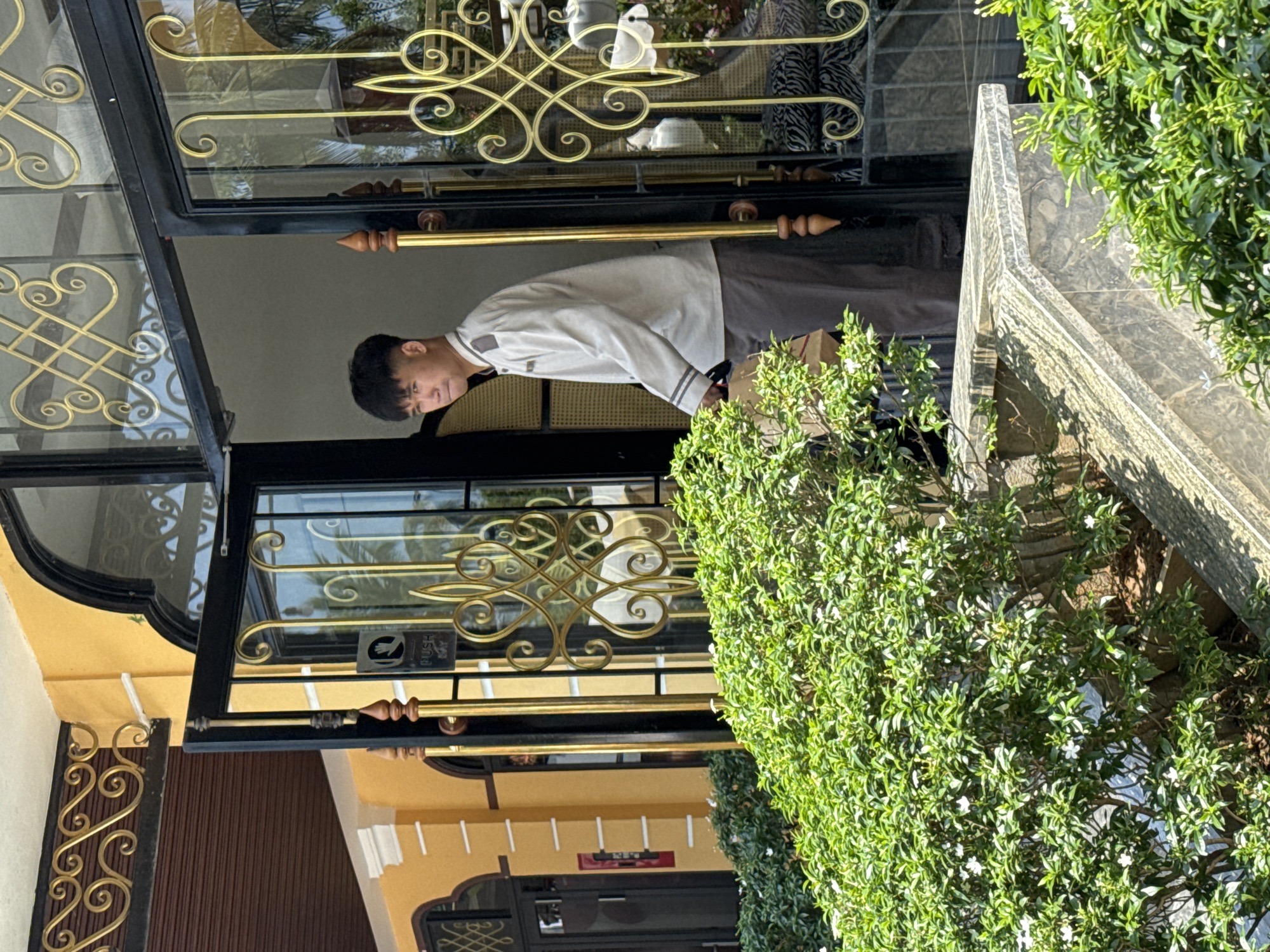 A male stands in the doorway of a building, holding onto the door handles with both hands, facing forward. The door has a decorative gold scrollwork design on the glass panels. The exterior of the building is yellow and brown. A plant with small white flowers grows on the right side of the door. The image was taken in Hội An Ward, Vietnam.