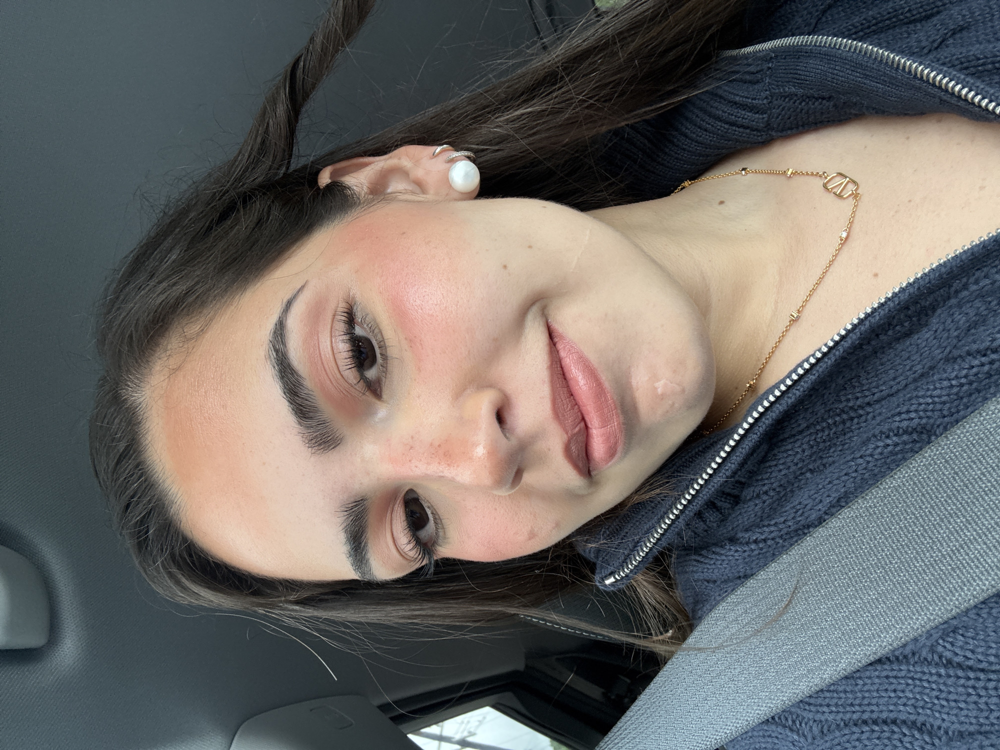 A woman is wearing a seatbelt and a Valentino necklace, possibly in a car. She has long brown hair, a pearl earring, and freckles. She appears to be wearing makeup.