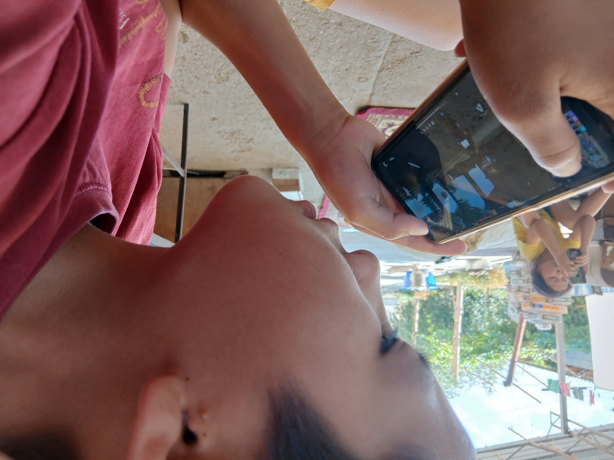 A close-up, profile shot shows a young person, viewed from the right side of their face, holding a smartphone horizontally in both hands and looking intently at the screen. The person wears a red t-shirt with a design of a yellow, outlined heart with a golden yellow stitching and pattern on the left chest area. The background is blurred but appears to be outdoors in a casual setting, possibly a yard or patio, with a shed-like structure overhead. In the further background, a younger person in a yellow t-shirt is visible, also looking at a smartphone. The ground appears to be a mix of concrete and earth with some scattered debris. The lighting suggests it is daytime. The image does not contain any specific landmarks or features that would allow for precise location identification within Kum-Aryk, Kyrgyzstan.