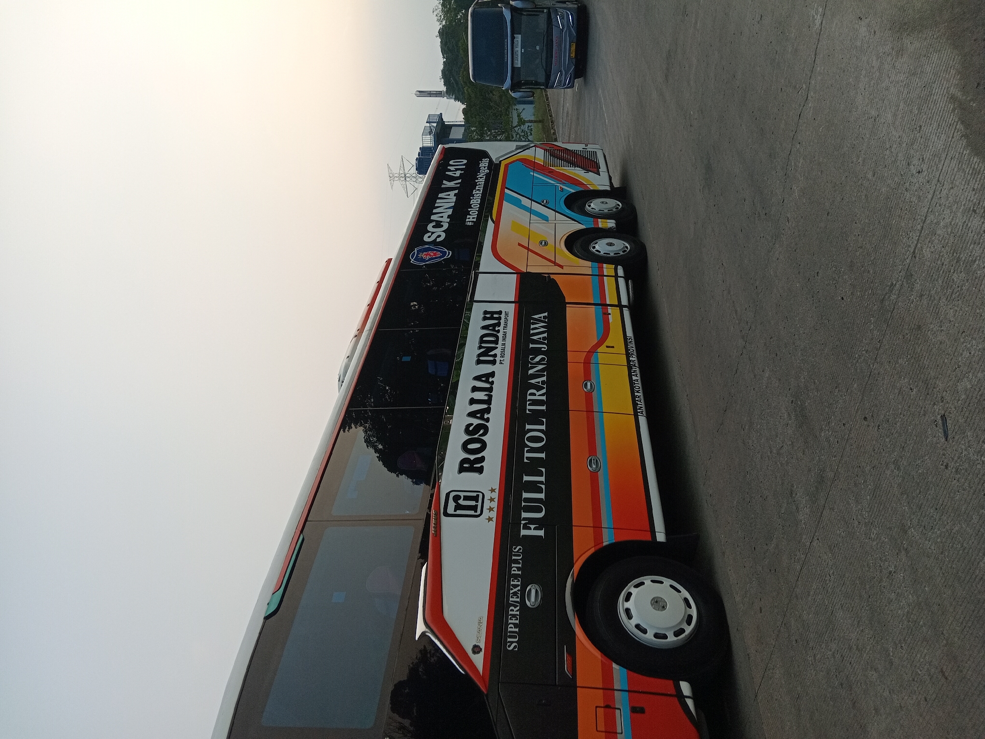The image shows two buses parked on an asphalt surface, possibly a bus station or parking lot. The bus in the foreground is a large, multi-colored bus with "ROSALIA INDAH FULL TOL TRANS JAWA" written on its side. It has a Scania K 410 logo and the hashtag #HoloBisEnakNgeBis. The bus in the background is a dark blue bus with "TRANS JAWA JAKARTA - YOGYA" written on it, and it also appears to be a passenger bus. The environment suggests an outdoor setting, possibly in the early morning or late evening due to the soft lighting and the sky's color. There are trees and power lines visible in the background. The presence of these buses implies a transportation hub. Without further information or visual cues, pinpointing the exact location within Depok, Indonesia, is not possible, but it is likely a major road or a transit point given the presence of inter-city buses.