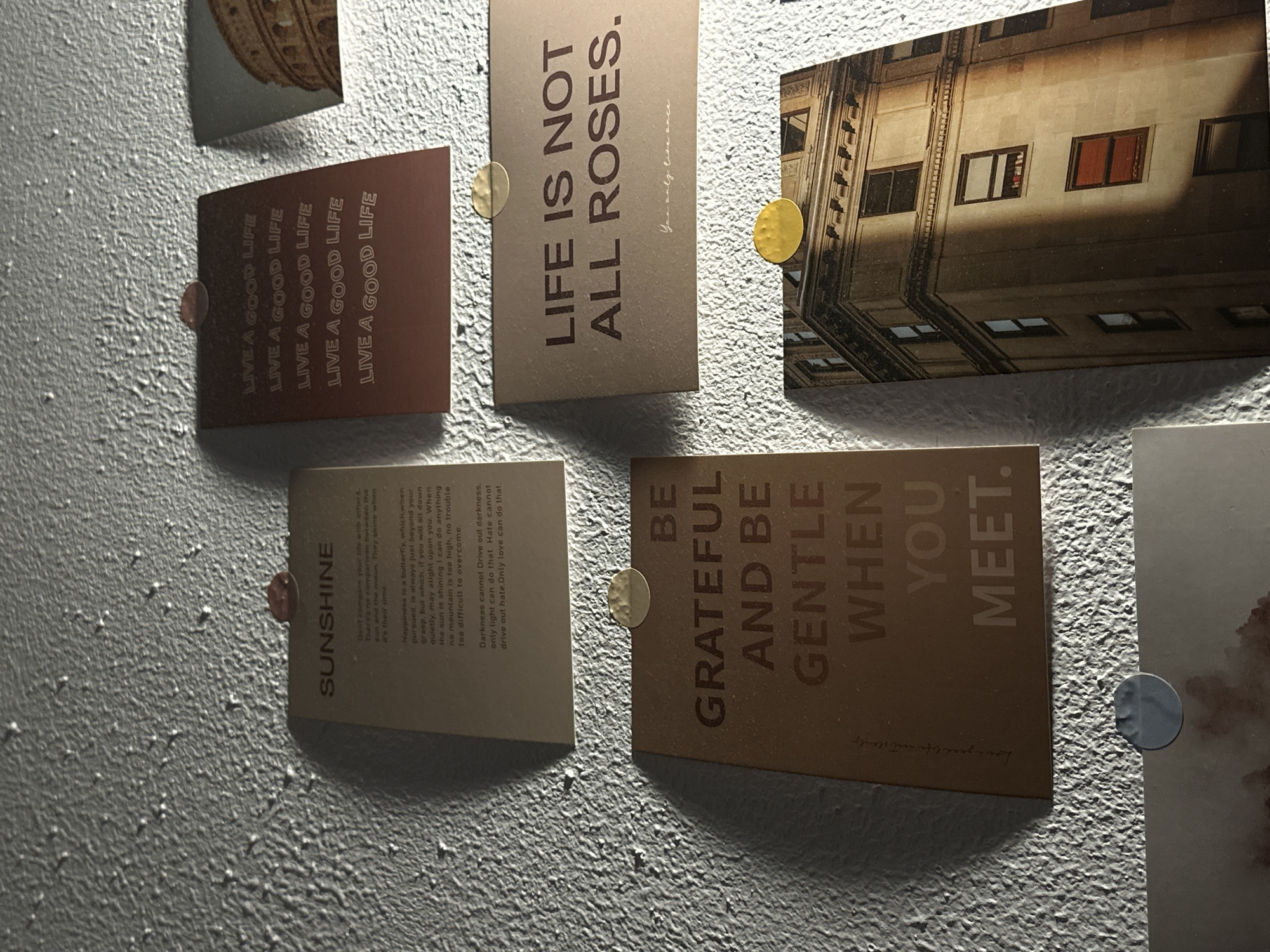 Several rectangular cards and one photo are displayed on a textured, light-colored wall, secured by small circular magnets or thumbtacks. The setting appears to be an indoor space under artificial lighting, casting subtle shadows.
One reddish-brown card prominently features the repeated phrase "LIVE A GOOD LIFE." Adjacent to it, a tan card reads "LIFE IS NOT ALL ROSES." with "You only live once" in a smaller script below. Above these, a sliver of another card with an architectural design is visible.
To the right, a photo card depicts the upper floors of an old building, viewed from a low angle, with numerous windows and a distinct diagonal shadow cast across its facade, indicating strong sunlight on the building itself.
Below the "LIVE A GOOD LIFE" card, another tan card is titled "SUNSHINE" and contains three inspirational quotes: "Don't compare your life with others. There's no comparison between the sun and the moon. They shine when it's their time", "Happiness is a butterfly, which when pursued, is always just beyond your grasp, but which, if you will sit down quietly, may alight upon you. When the sun is shining I can do anything no mountain is too high, no trouble too difficult to overcome.", and "Darkness cannot Drive out darkness, only light can do that. Hate cannot drive out hate. Only love can do that."
Next to the "SUNSHINE" card, a brown card advises, "BE GRATEFUL AND BE GENTLE WHEN YOU MEET." followed by a small cursive phrase "Live your best life." A partially visible card with indeterminate content is seen at the bottom right corner. The fasteners holding the cards vary in color, including gold, reddish-brown, and silver.