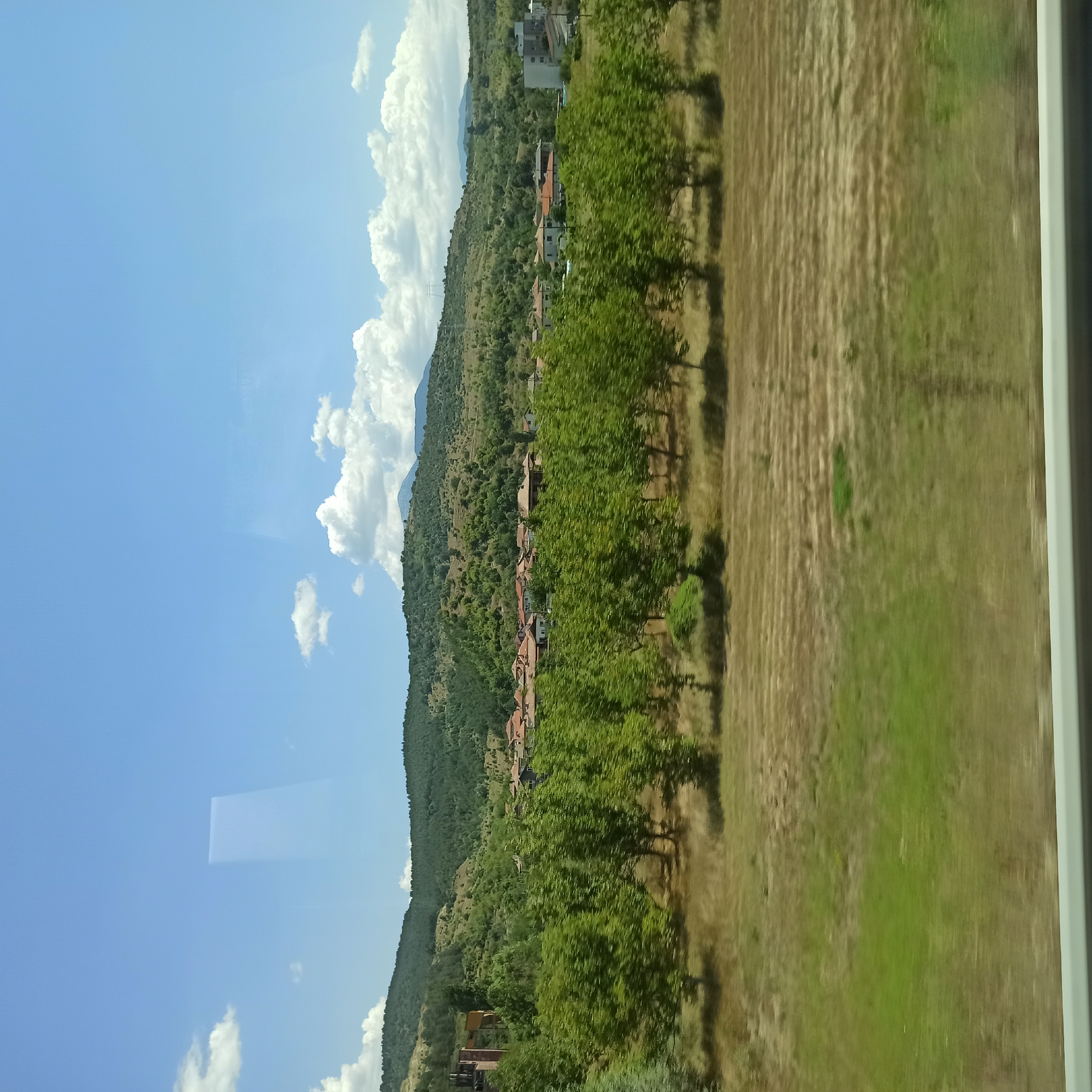 The image captures a rural landscape with a small village nestled in a valley. The foreground consists of a dry, mowed field with distinct rows of stubble, giving way to a line of green trees in the middle ground. Beyond the trees, a village with red-tiled roofs and some modern buildings is visible, set against a backdrop of lush, green, rolling hills. The sky above is a vibrant blue, dotted with fluffy white clouds. The scene appears to be taken from a moving vehicle, indicated by the slight blur in the foreground vegetation and the reflection on the surface, likely a window. The overall impression is one of a peaceful, sunny day in a Bulgarian countryside, potentially near the town of Simitli.