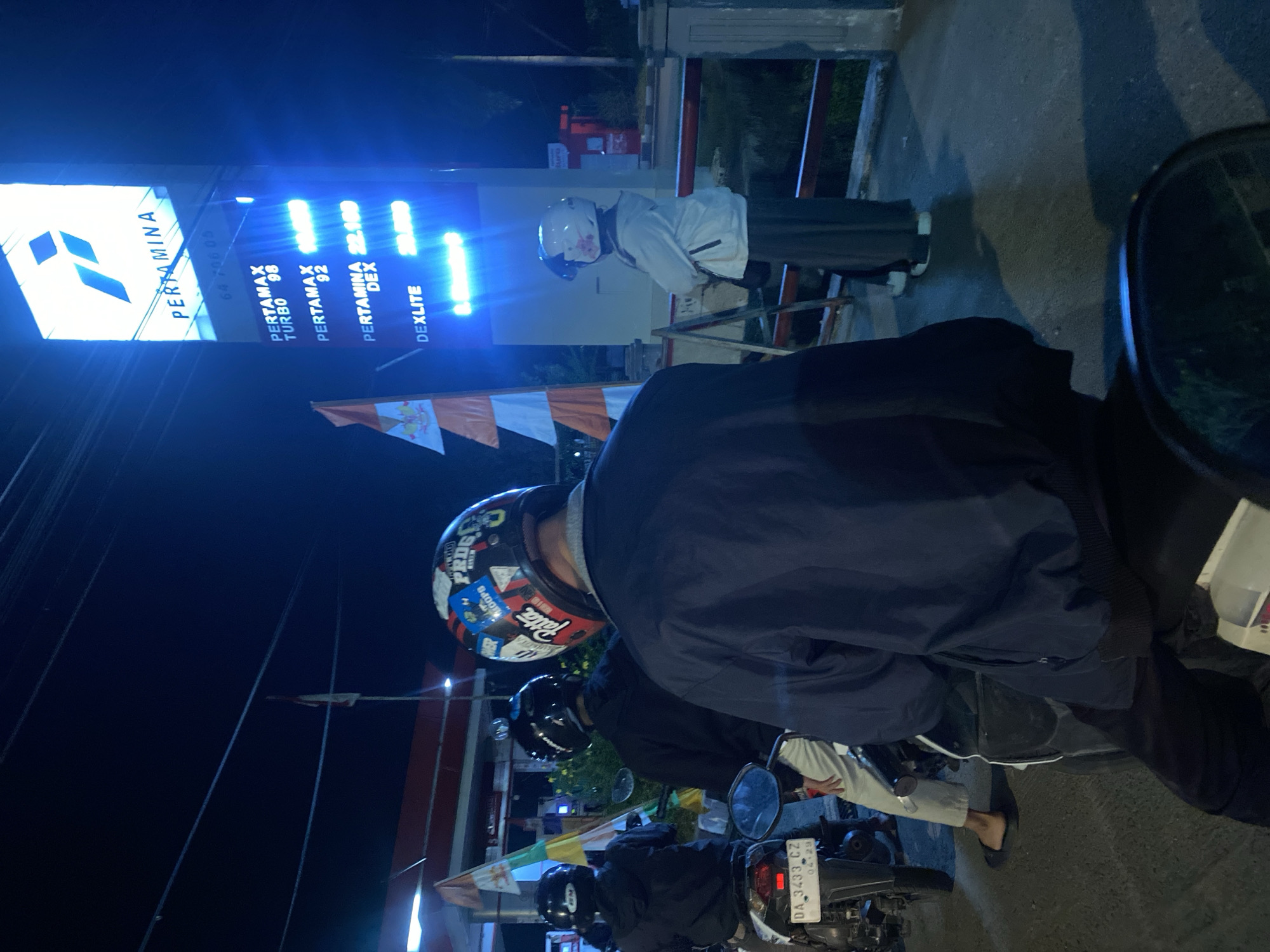 The image captures a nighttime scene at a Pertamina gas station in Gambut Barat, Indonesia. The main focus is a tall, illuminated Pertamina sign displaying fuel prices. The sign shows "PERTAMAX TURBO 98" with a price of "16.000", "PERTAMAX 92" priced at "14.500", "PERTAMINA DEX" at "22.100", and "DEXLITE" at "20.000". In the foreground, three individuals on motorcycles are visible. The person in the center, wearing a black jacket and white pants, is partially obscured. To the left, another individual in a black jacket is seated on a motorcycle with license plate "DA 3433 CZ". The person on the right, seen from the back, is wearing a dark jacket and a helmet adorned with multiple stickers, including one that says "Patta". A fourth individual, wearing a white jacket and a pink-flowered helmet, stands to the right, facing the gas station. The environment appears to be an outdoor gas station with a canopy visible in the background, along with some lights and flags. The ground is wet, suggesting recent rain.