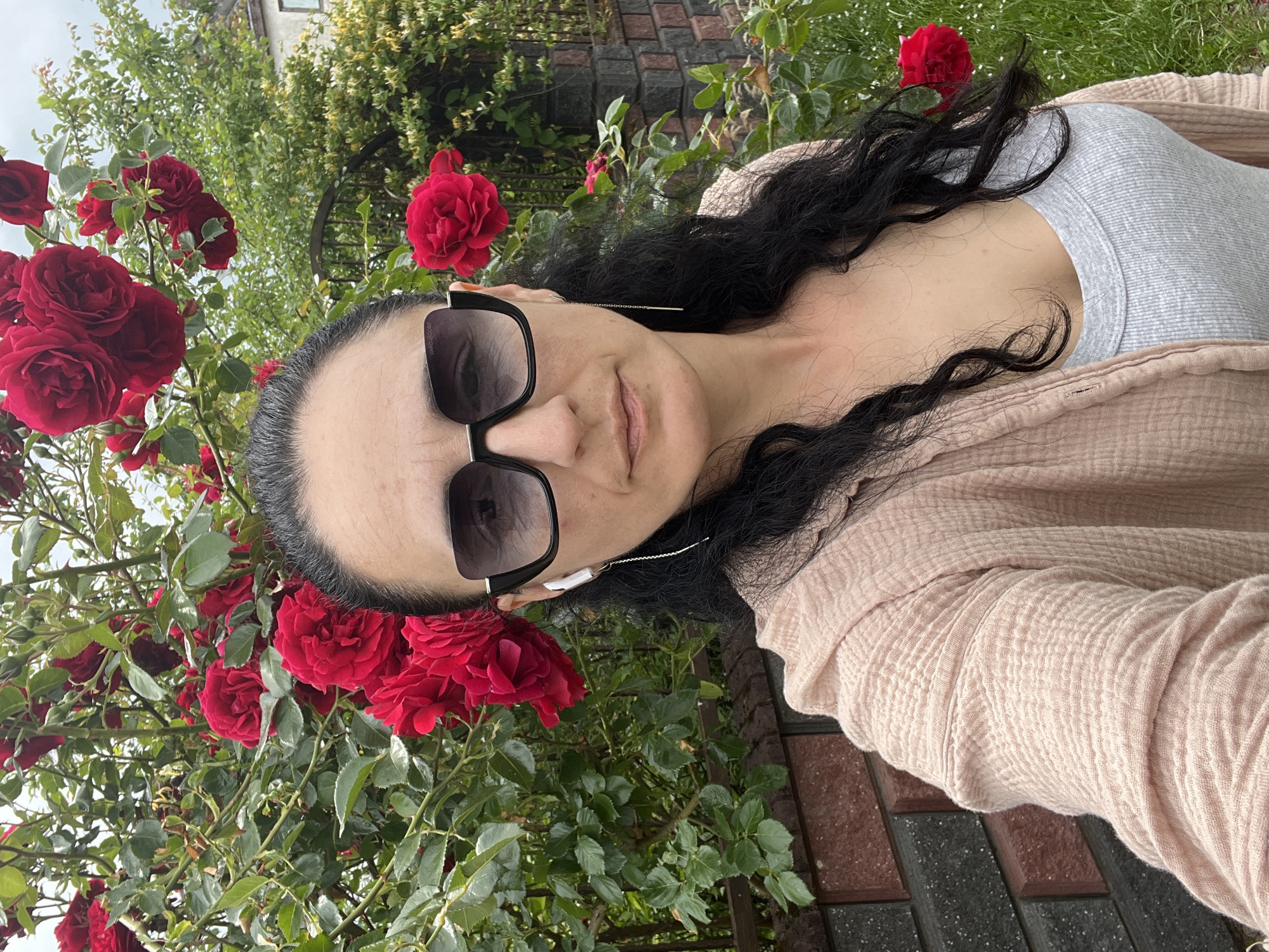 This image is a selfie-style portrait, rotated 90 degrees clockwise, featuring a woman outdoors in what appears to be a garden setting in Onokivtsi, Ukraine.
The primary subject is a woman with long, dark, wavy hair. She is wearing large, black-framed sunglasses that cover her eyes, with the text "MARC JACOBS" visible on the left arm. Her expression is neutral, with her lips slightly parted. She has fair skin and is dressed in a light grey ribbed top, layered with a light pink or beige crinkled-texture cardigan. White wired earbuds are visible, with the wire running down her side.
She is positioned in front of a vibrant rose bush laden with numerous dark red roses and lush green foliage, which prominently frames the left side of the image. The background reveals a paved area made of dark grey and reddish-brown rectangular pavers. Further back, dense green trees and bushes are visible, along with a partial view of a building or house with a window. The natural lighting suggests it is daytime, likely under bright, diffused light as there are no harsh shadows. The scene conveys a relaxed outdoor moment amidst blooming flowers.